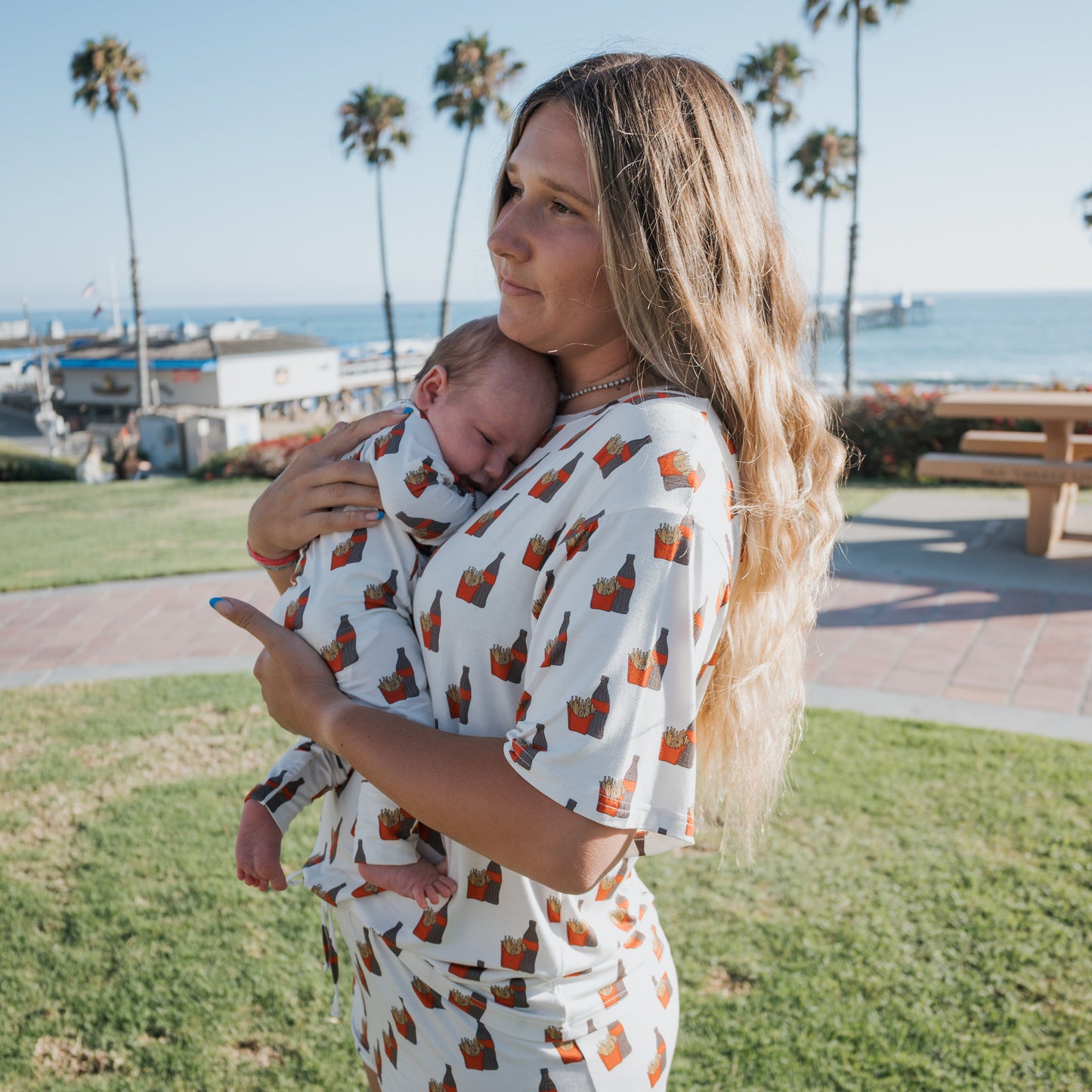 A woman with long blonde hair holds a sleeping baby by the beach. Both wear matching forever french baby Bamboo Zip Pajamas | Fries & Cola, gentle on sensitive skin. Palm trees, grass, benches, and ocean create a tranquil scene.