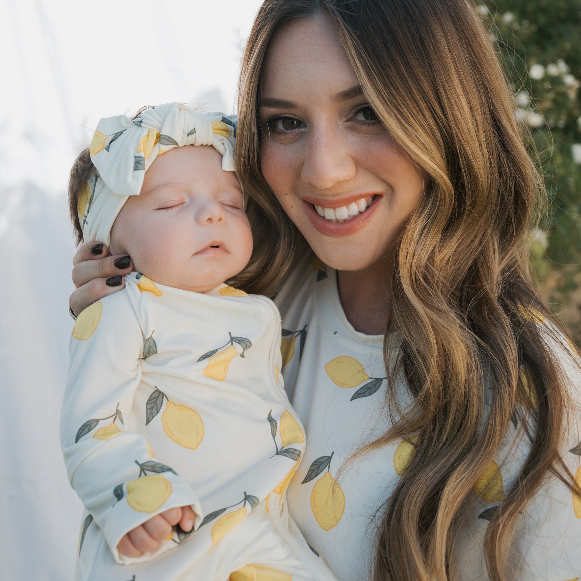 A smiling woman holds a sleeping baby outdoors, both dressed in matching white Bamboo Zip Pajamas | Lemon by forever french baby with yellow lemon patterns. The baby also wears a matching headband. They're surrounded by greenery.