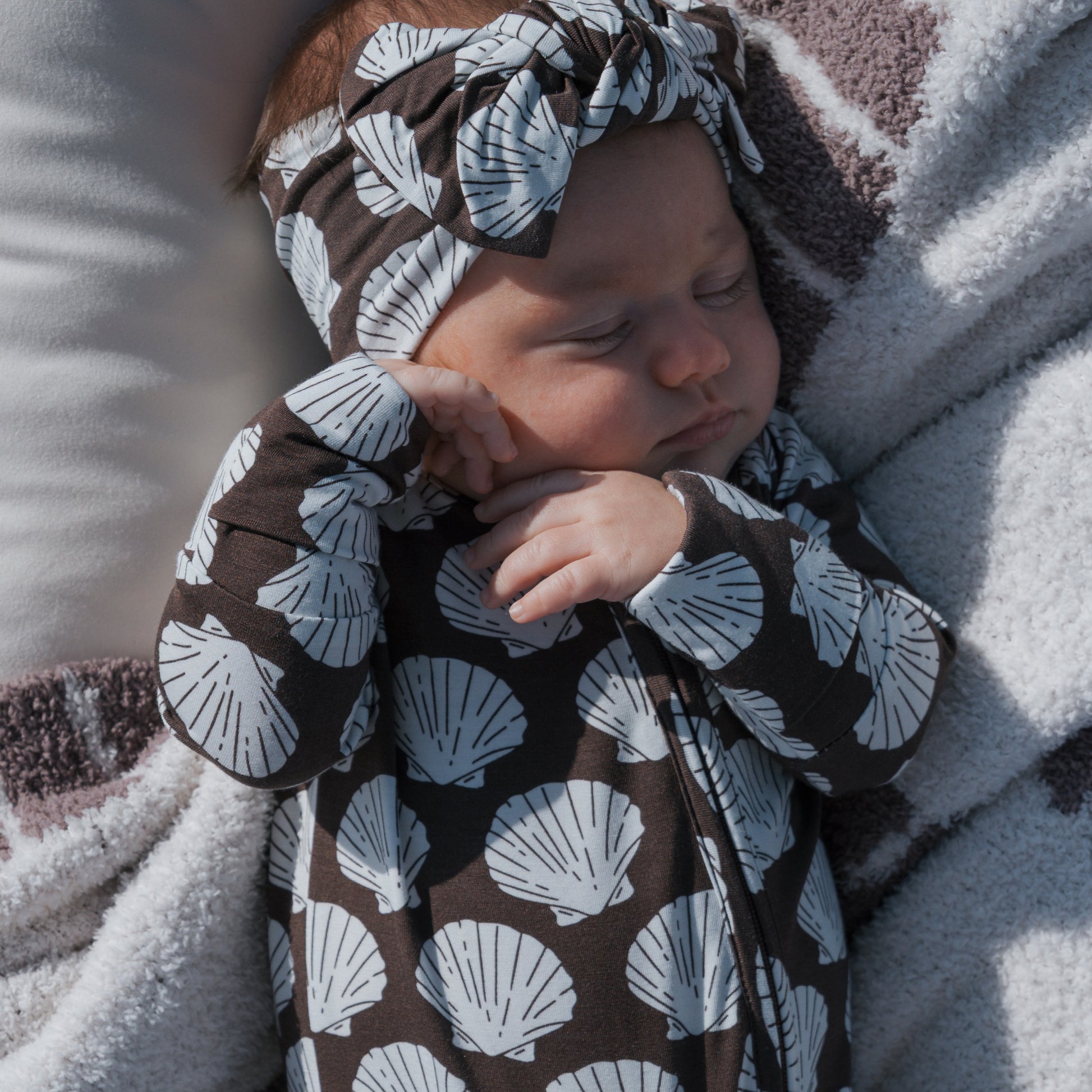 A baby sleeps soundly in forever french baby's Bamboo Zip Pajamas | Seashells, featuring breathable, soft bamboo fabric and a matching headband with white seashells on a dark background for ultimate comfort.