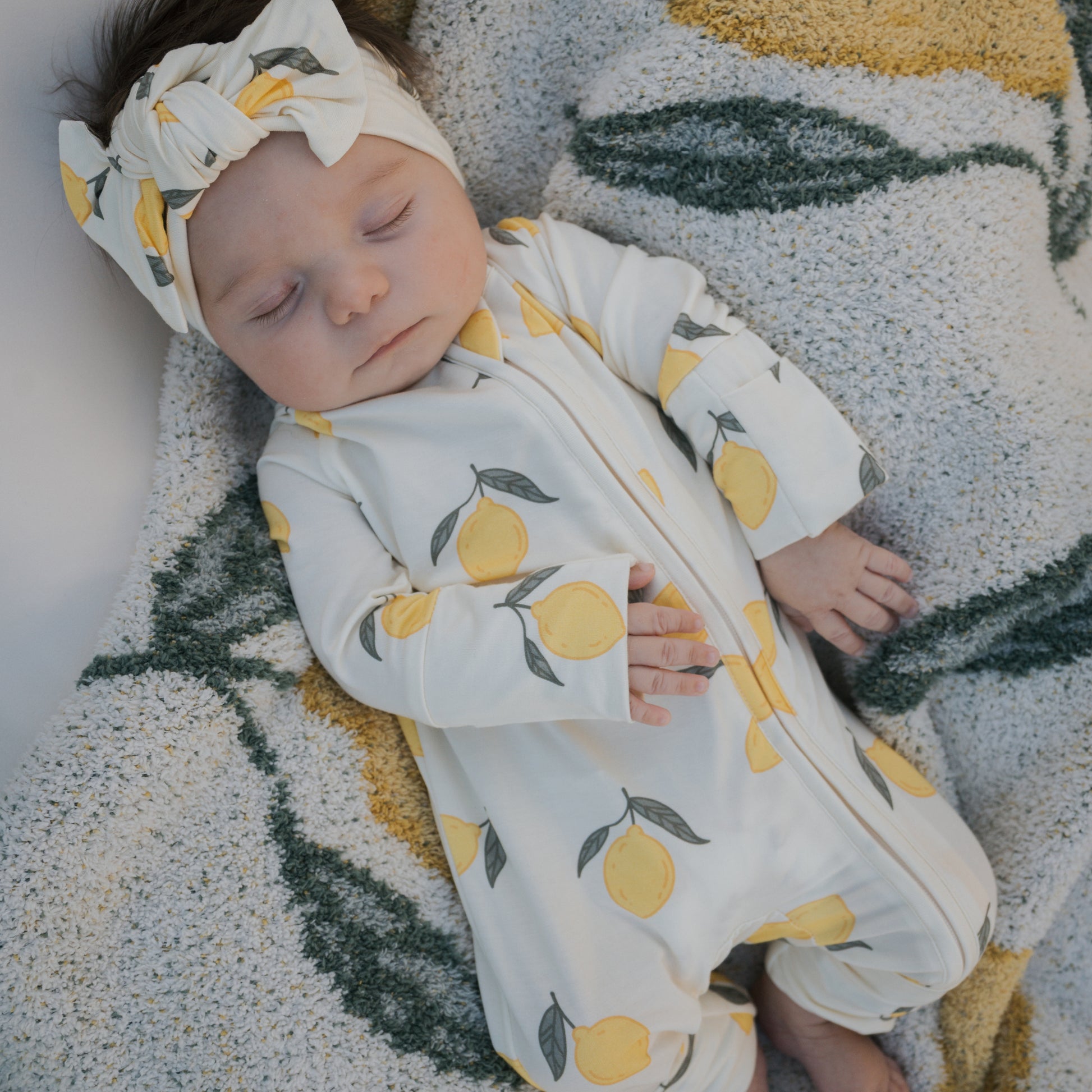 A baby sleeps on a soft, textured blanket, wearing forever french baby Bamboo Zip Pajamas | Lemon—a breathable white onesie with a headband featuring yellow lemons and green leaves.