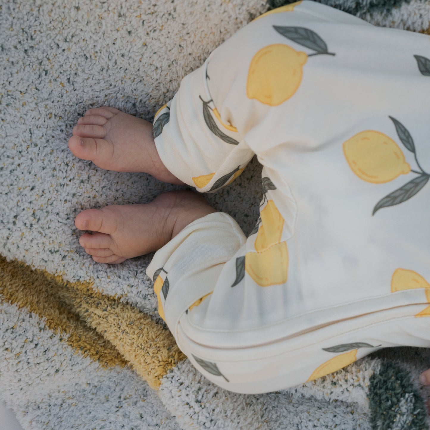 A close-up of a baby’s legs and feet resting on a textured blanket, wearing forever french baby Bamboo Zip Pajamas | Lemon—hypoallergenic white pants with a charming lemon and leaf pattern.