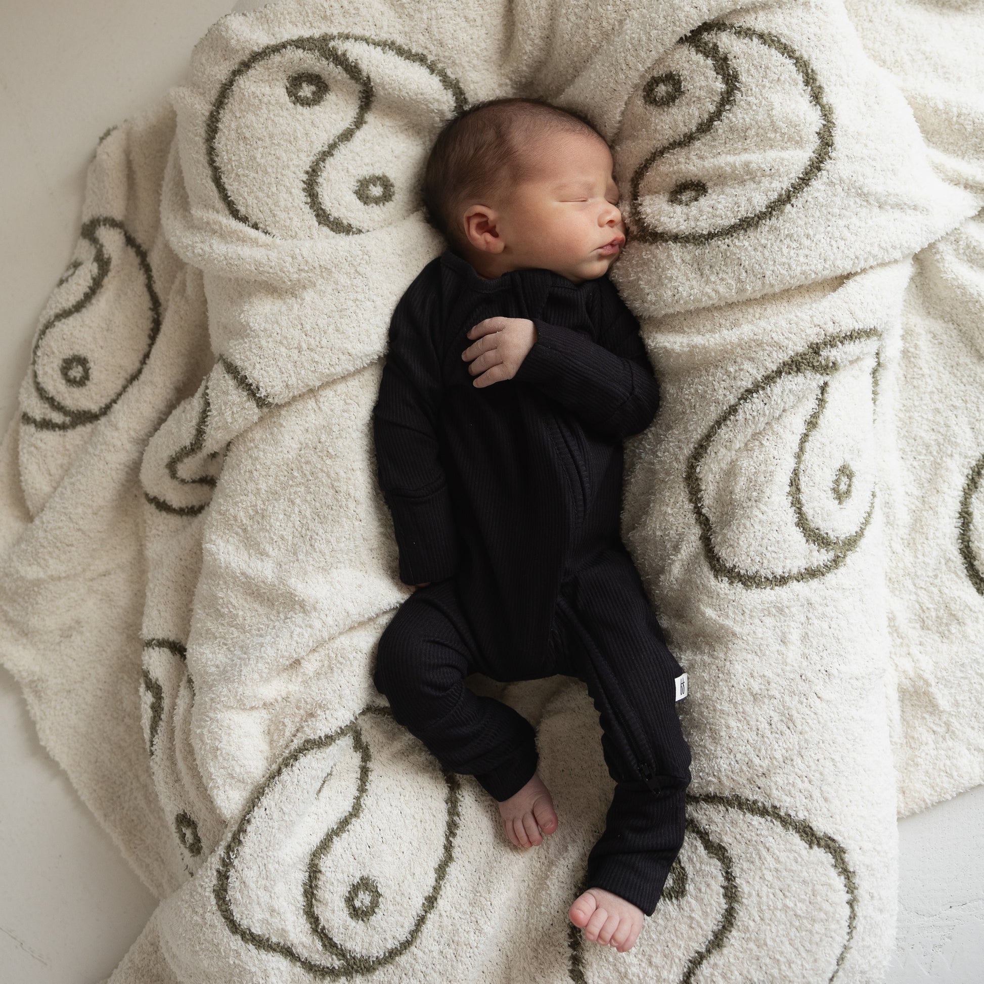 A newborn sleeps peacefully in forever french baby's Bamboo Zip Pajamas | Ribbed Black on a fluffy white blanket with large green yin-yang symbols.