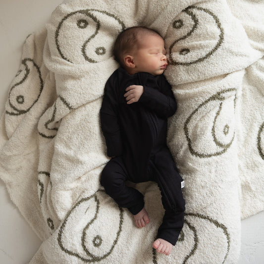 A newborn sleeps peacefully in forever french baby's Bamboo Zip Pajamas | Ribbed Black on a fluffy white blanket with large green yin-yang symbols.