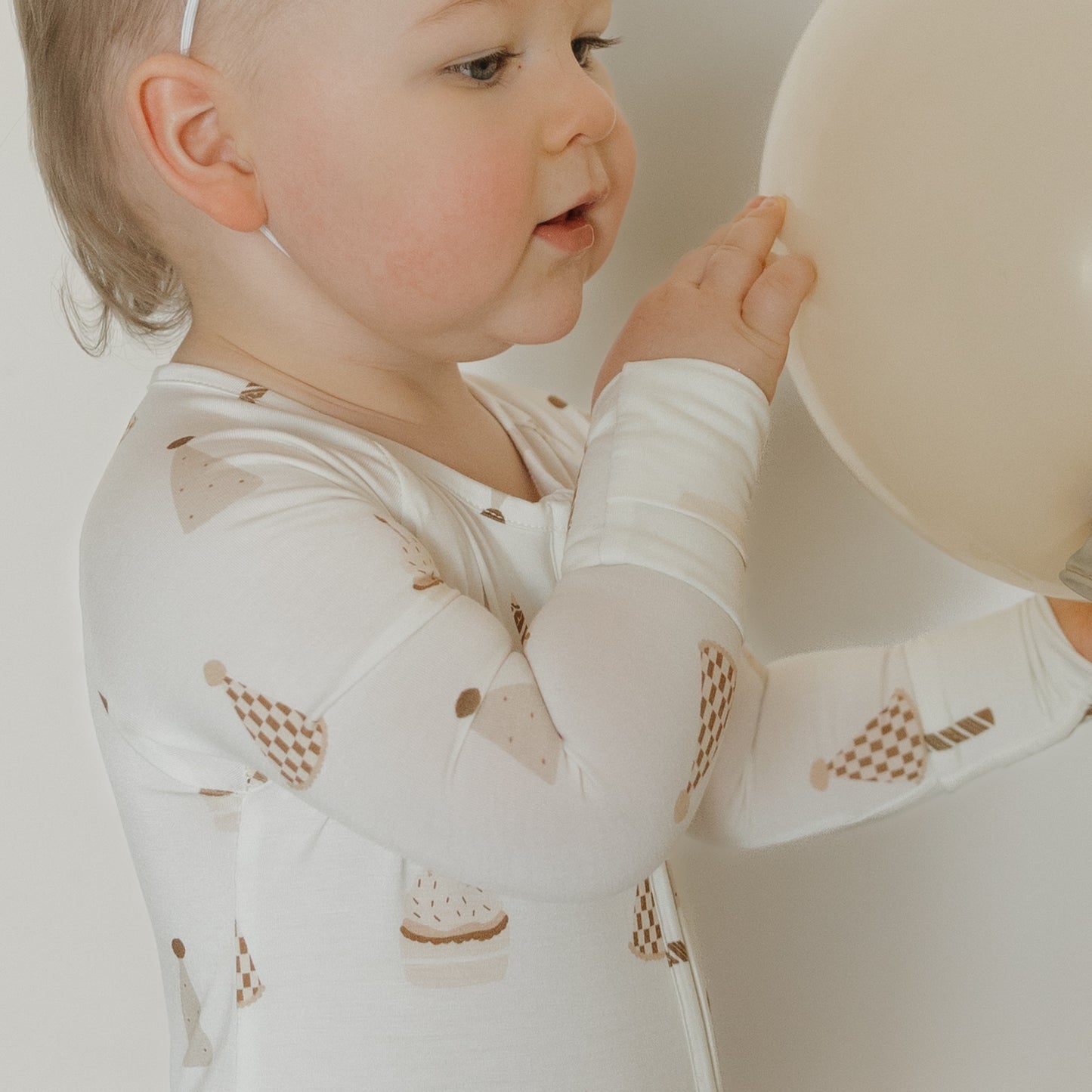 A toddler dressed in the forever french baby Bamboo Zip Pajamas | Birthday Boy—featuring festive party hat patterns on soft, breathable fabric—touches a large beige balloon while standing against a plain light backdrop.
