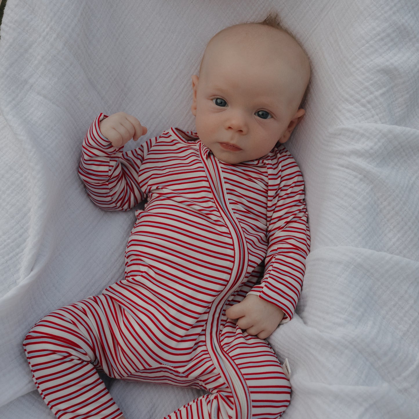 A baby in forever french baby's Bamboo Zip Pajamas | Heritage Stripe—a red and white striped, breathable sleepwear—lies on a soft white blanket, gazing up calmly.
