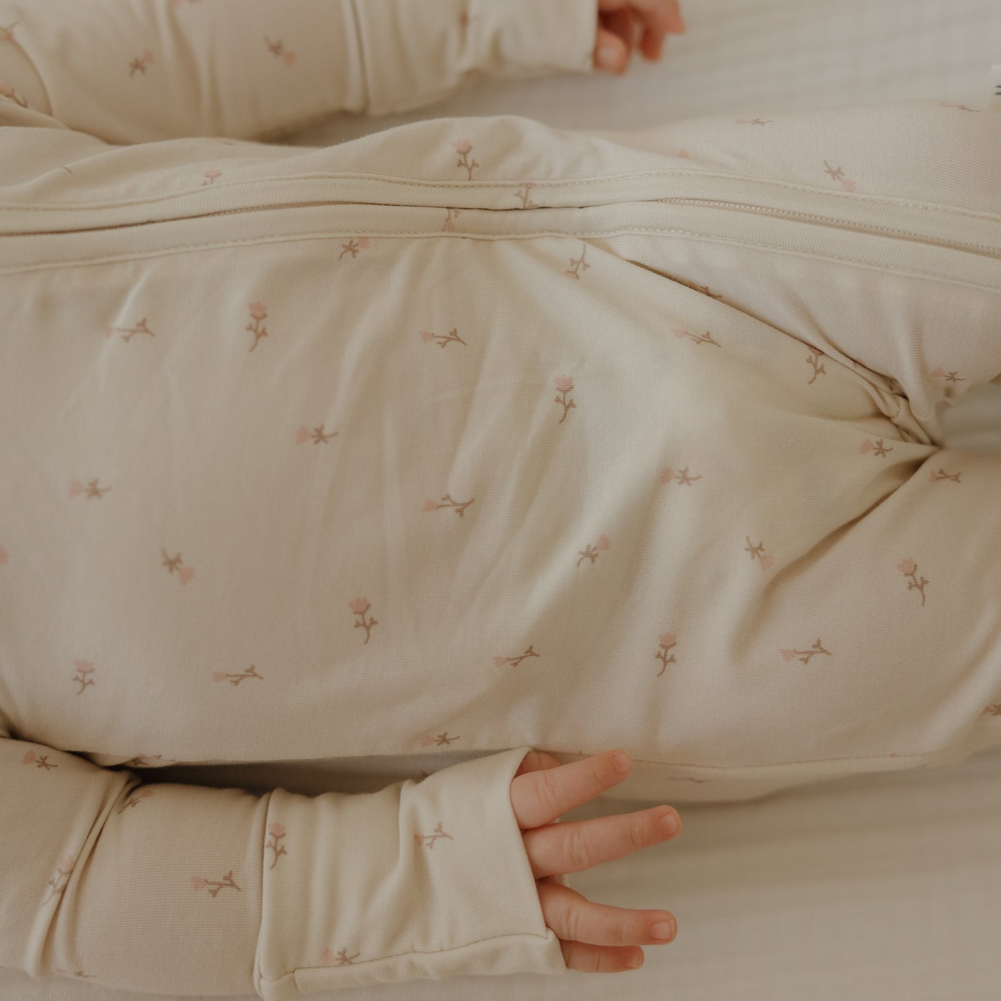 A close-up of a baby lying on a light surface in forever french baby’s Bamboo Zip Pajamas | Lucy—breathable cream-colored sleepwear with a subtle floral pattern. Only the baby's hands and part of their legs and arms are visible.