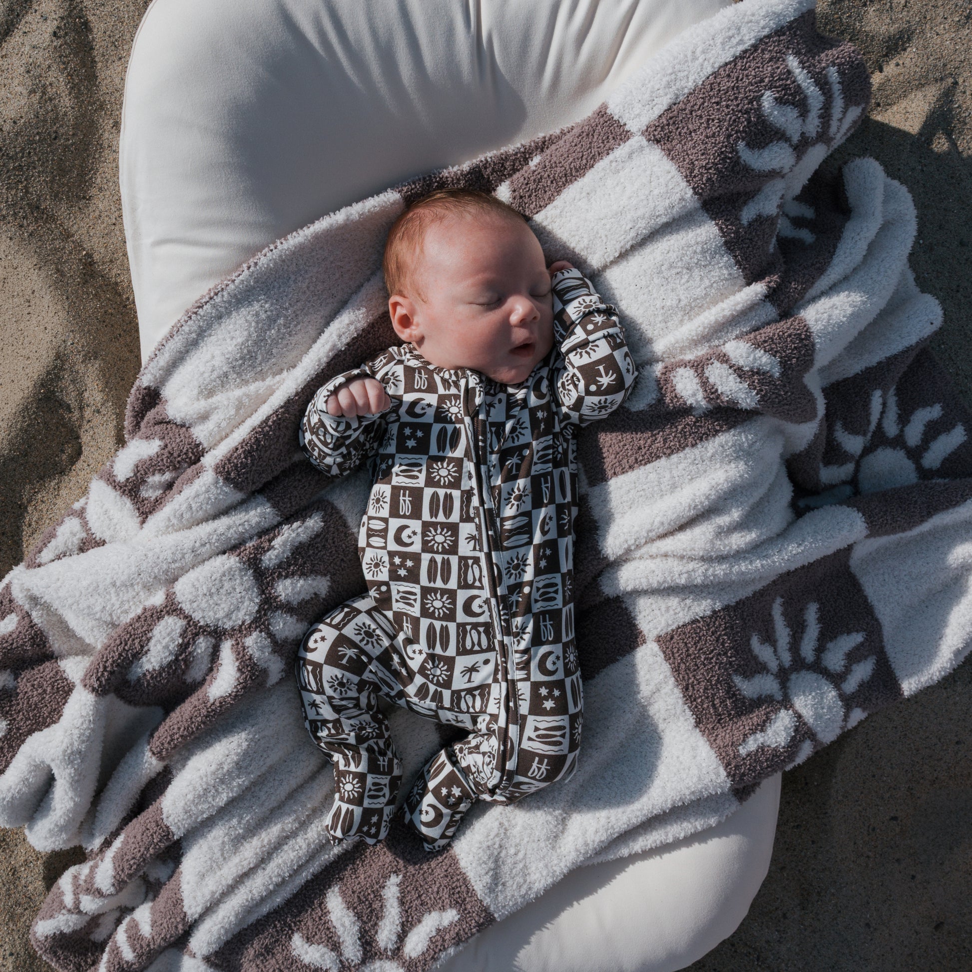 A baby in forever french baby's Bamboo Zip Pajamas | Summer Check lies on a white cushion, wrapped in a brown and white paw print blanket, resting on sandy ground.