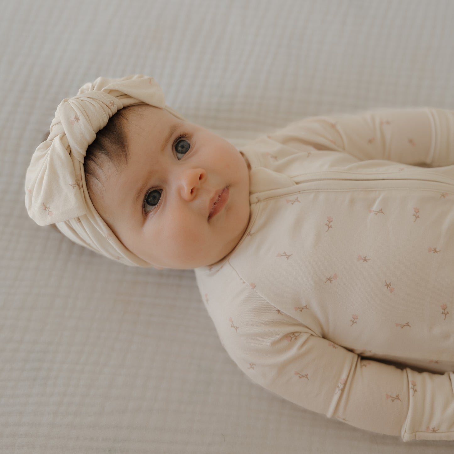 A baby in forever french baby's Bamboo Zip Pajamas | Lucy with a matching knotted headband rests on a soft, textured surface, gazing upward with a calm expression.