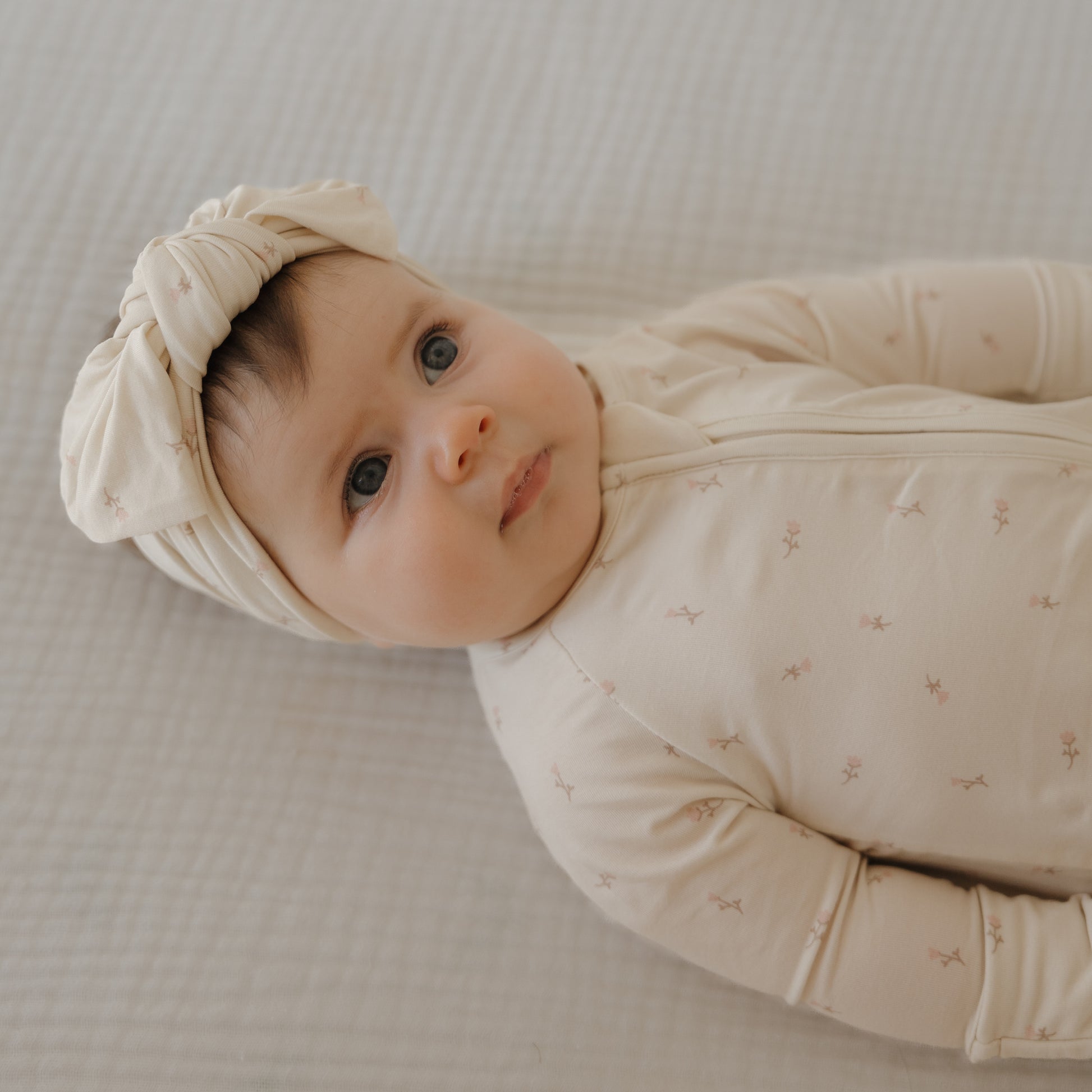 A baby in forever french baby's Bamboo Zip Pajamas | Lucy with a matching knotted headband rests on a soft, textured surface, gazing upward with a calm expression.