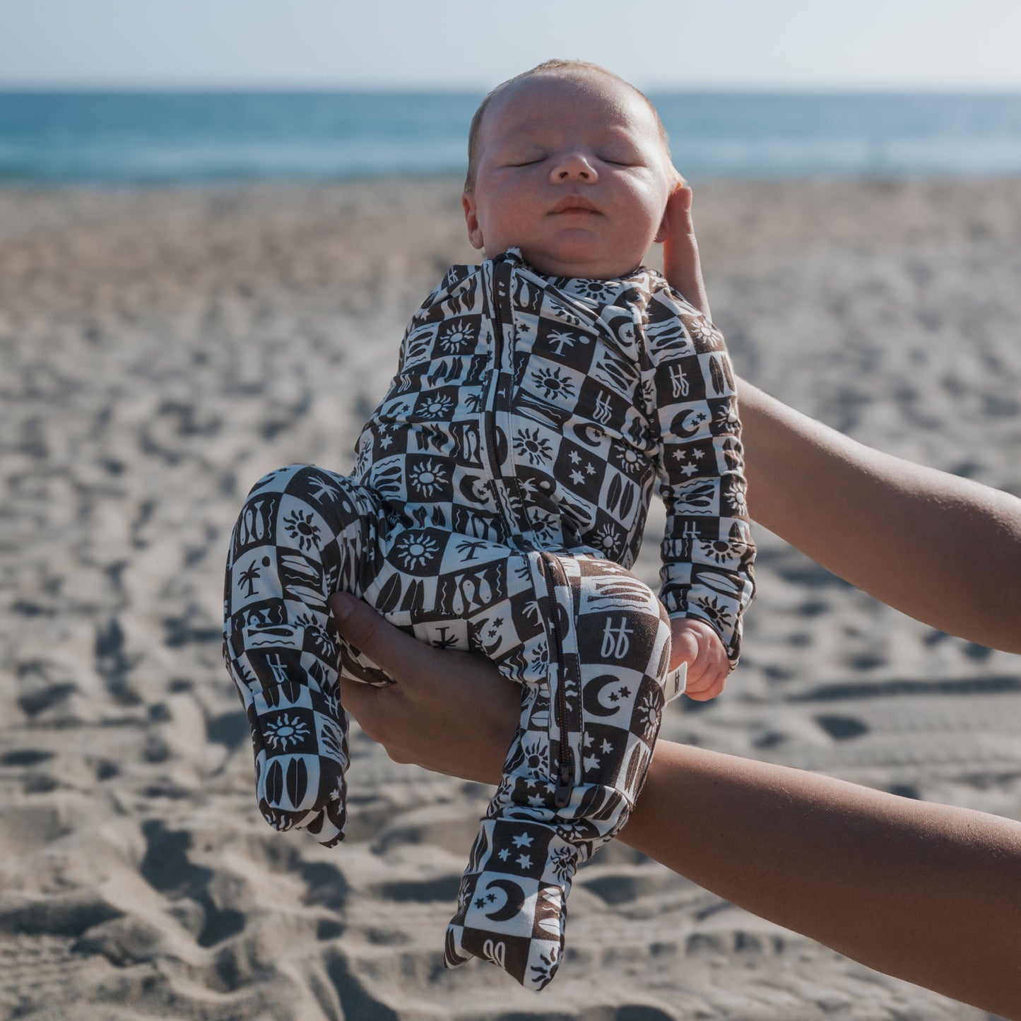 A newborn in forever french baby Bamboo Zip Pajamas | Summer Check is cradled in adult hands on a sandy beach, with the sea and sky in the background.
