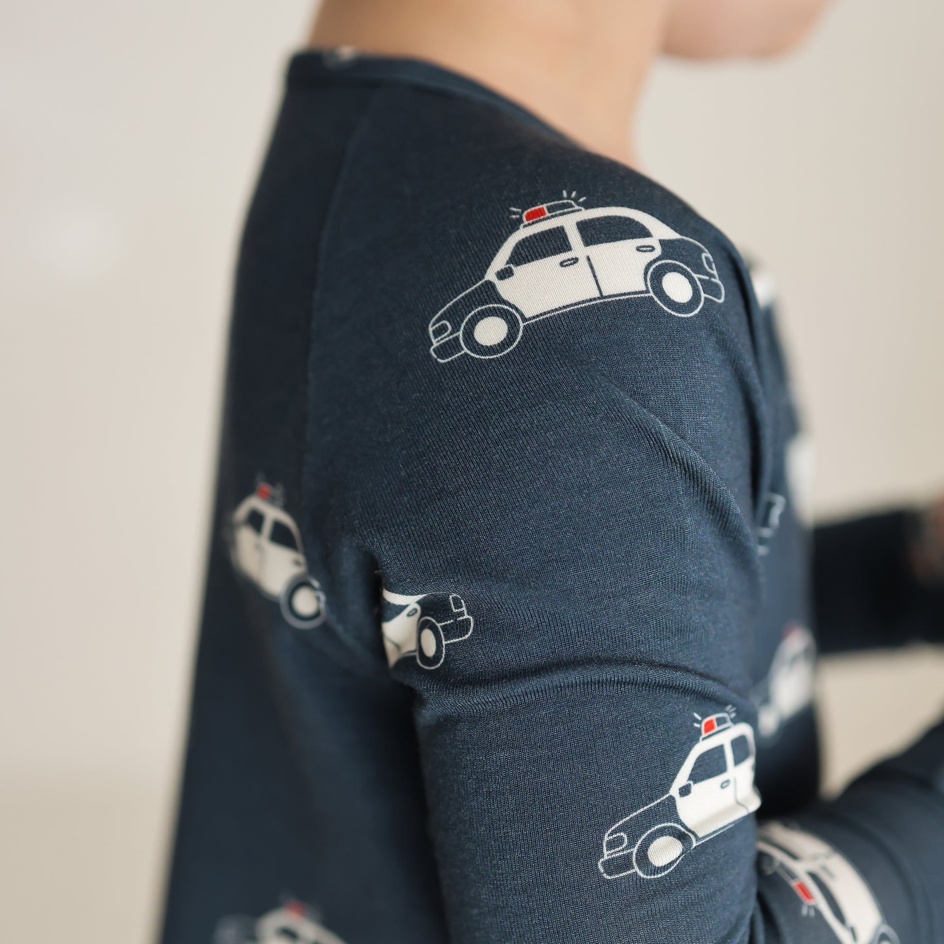 A child wears forever french baby Bamboo Zip Pajamas | Police Cruiser—dark blue sleepwear patterned with small white police cars and red lights, shown from upper body to shoulder against a softly blurred background.