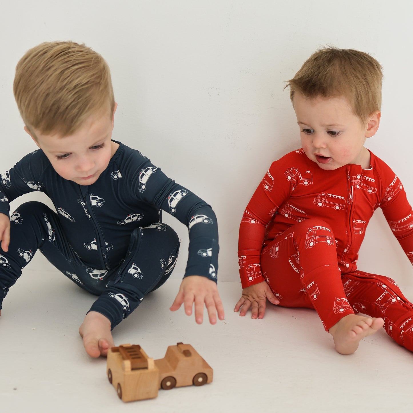 Two young kids sit on the floor in forever french baby Bamboo Zip Pajamas—one in navy, one in the Fire Truck print. The child in blue reaches for a toy truck, both cozy and comfortable in soft, breathable sleepwear.