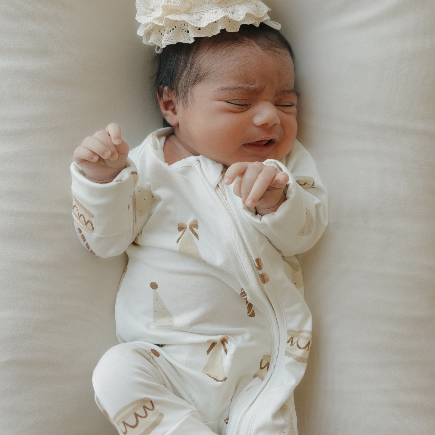 A newborn baby wearing forever french baby's Bamboo Zip Pajamas | Birthday Girl—a soft, white onesie adorned with light brown patterns and paired with a lace headband—lies on a white surface, appearing to cry or fuss.