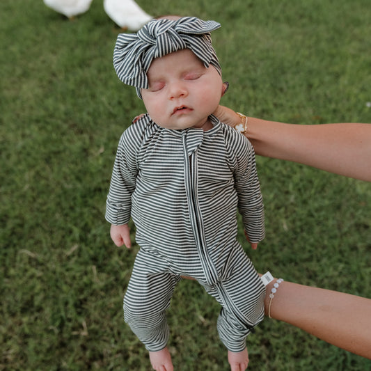 A baby dressed in forever french baby’s Bamboo Zip Pajamas in Forest Stripe Ribbed and a black and white striped headband is sleeping while being held upright outdoors on green grass.