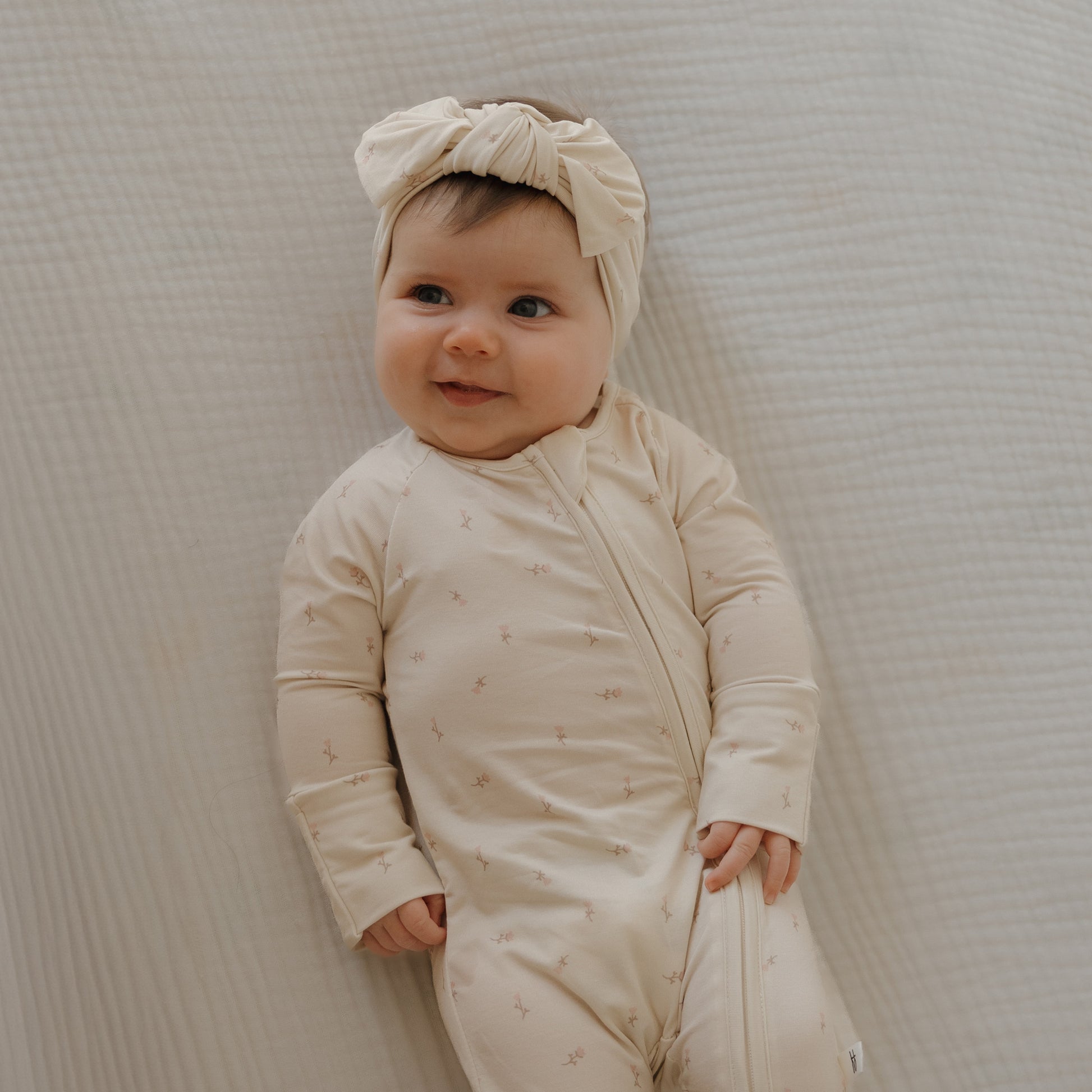 A smiling baby wears forever french baby’s Bamboo Zip Pajamas | Lucy—a light beige onesie with subtle patterns and a matching headband—while lying on a textured cream-colored blanket.