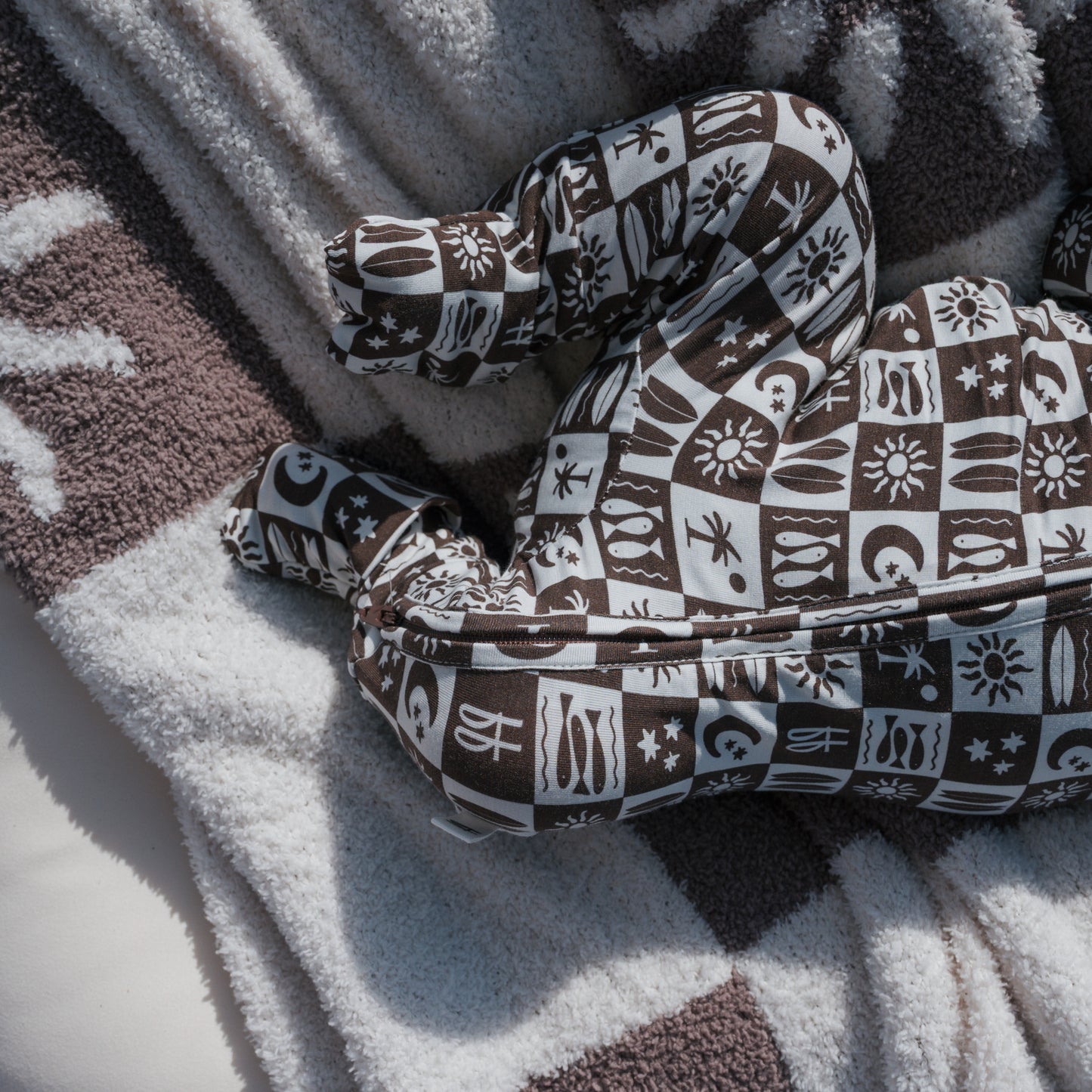 A brown and white patterned pillow shaped like legs rests on a textured, striped blanket, evoking the soft comfort of forever french baby's Bamboo Zip Pajamas | Summer Check as sunlight casts gentle shadows over the cozy scene.