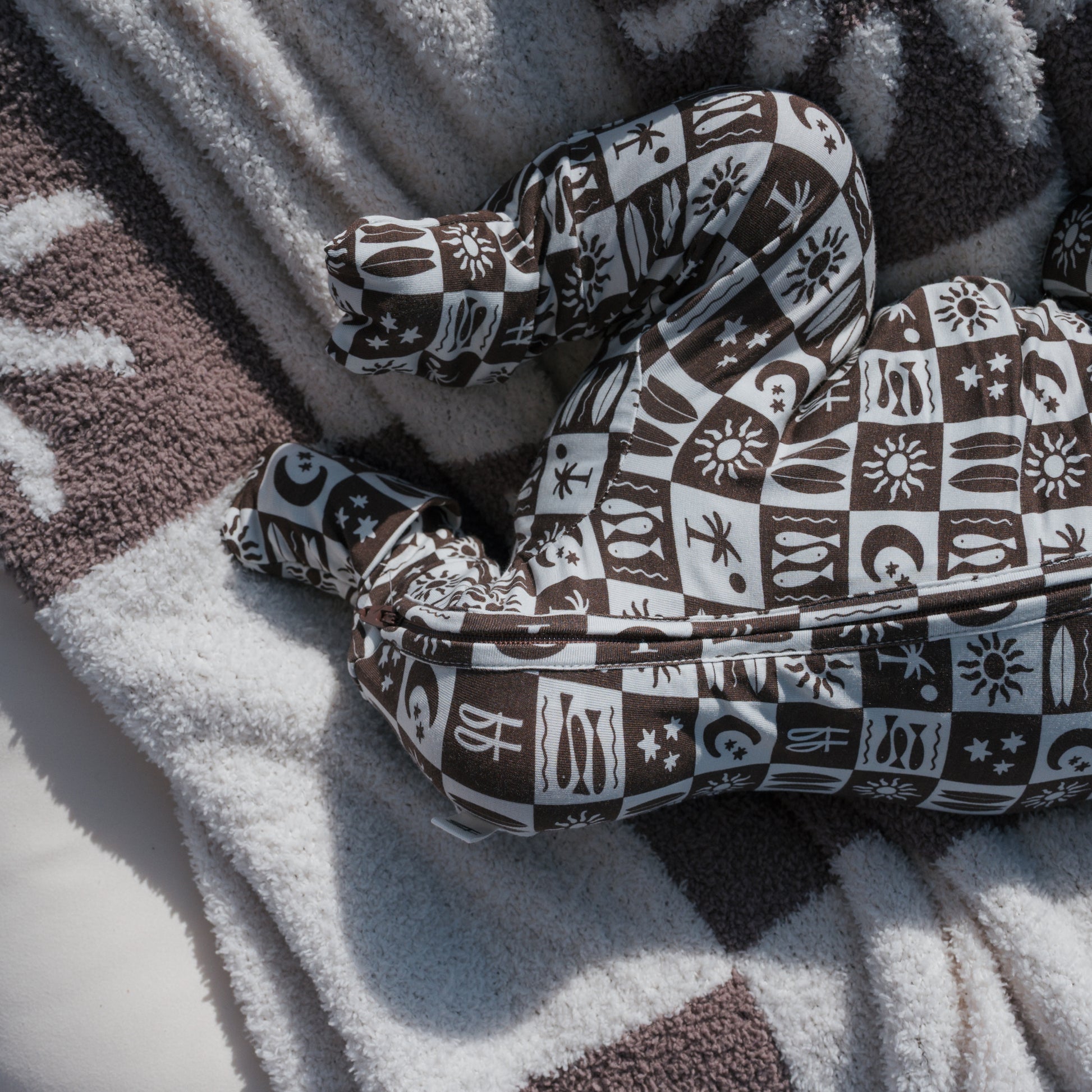 A brown and white patterned pillow shaped like legs rests on a textured, striped blanket, evoking the soft comfort of forever french baby's Bamboo Zip Pajamas | Summer Check as sunlight casts gentle shadows over the cozy scene.