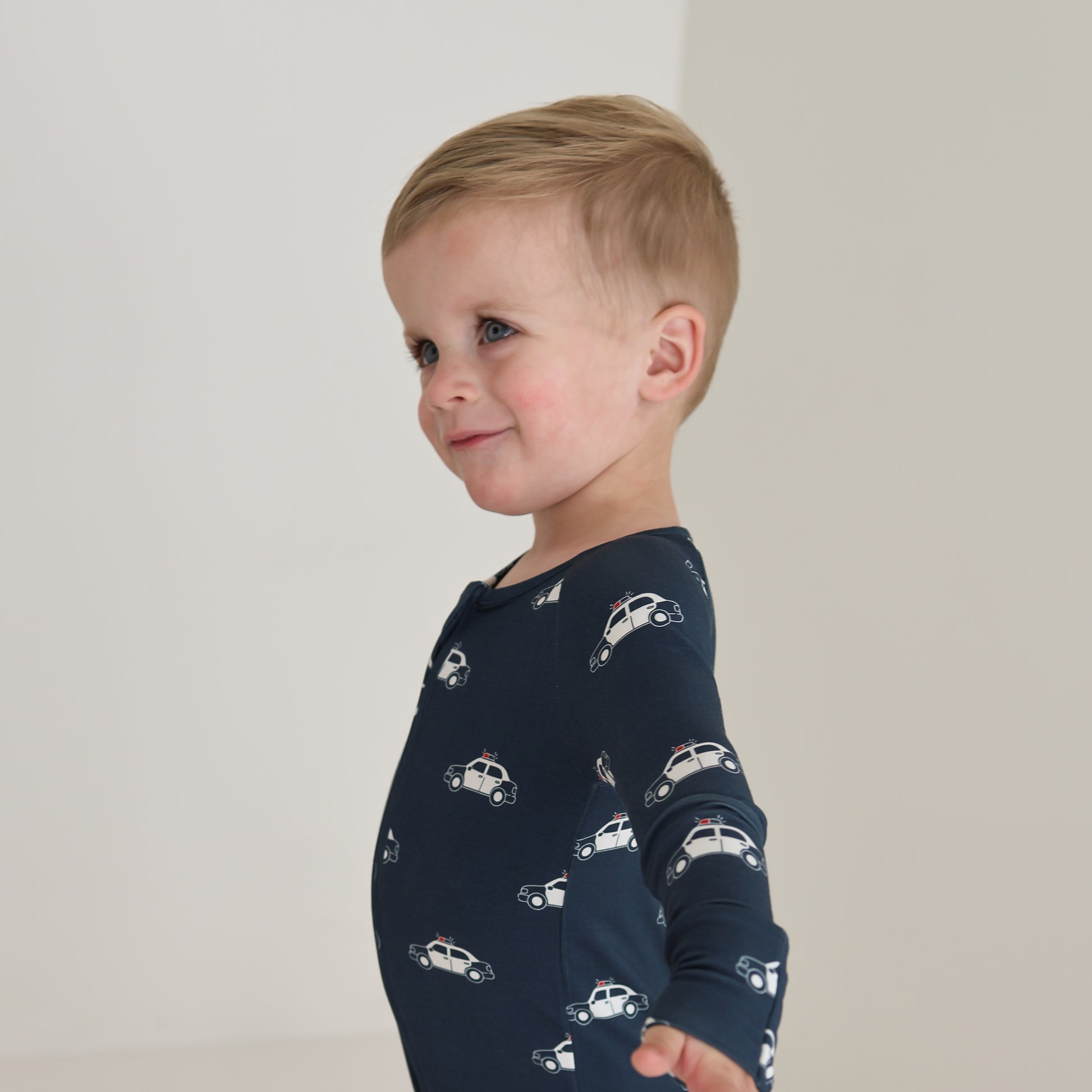 A young child with short blonde hair smiles sideways with arms outstretched, wearing forever french baby Bamboo Zip Pajamas | Police Cruiser—dark blue breathable sleepwear featuring white police car patterns against a light background.