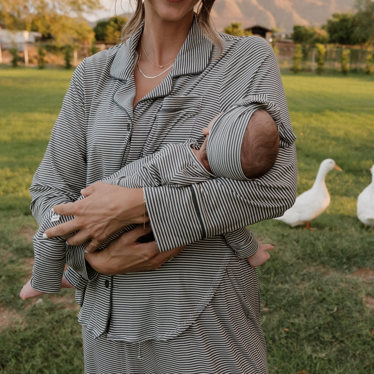 A woman in striped pajamas stands on the grass holding a baby dressed in matching stripes, wearing the forever french baby Bamboo Head Wrap | Forest Stripe Ribbed. Two white ducks are visible near a fence and trees in the background.