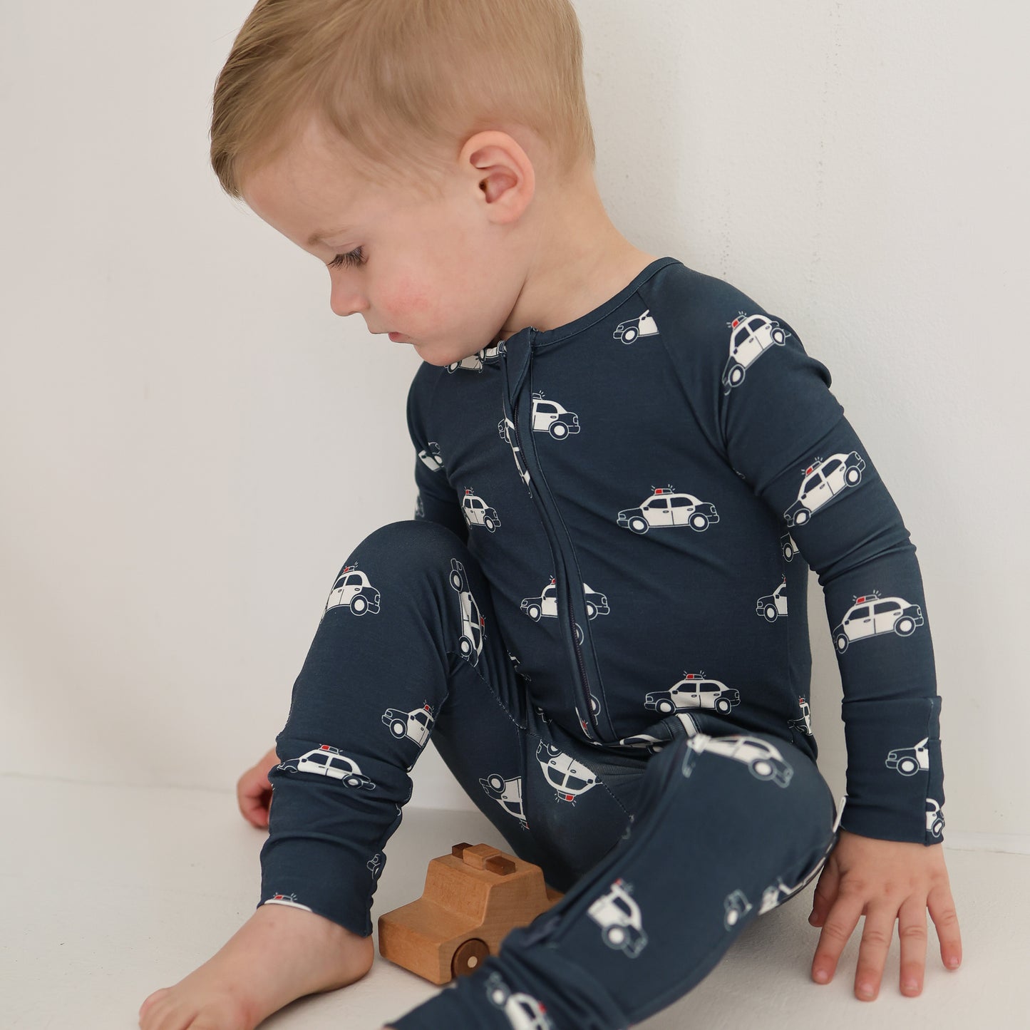 A young child sits on the floor in forever french baby Bamboo Zip Pajamas | Police Cruiser, featuring breathable bamboo fabric and fun police car prints, holding a wooden toy car while gazing down and to the side.