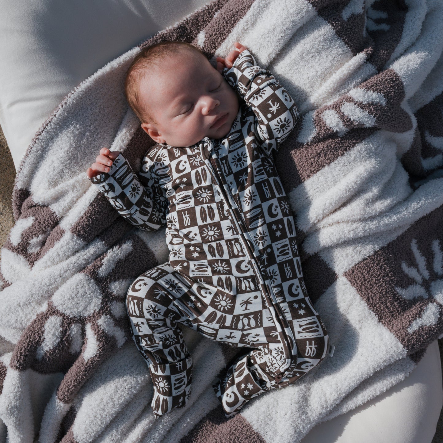 A baby sleeps soundly on a cozy blanket, dressed in forever french baby Bamboo Zip Pajamas in Summer Check, with one tiny arm resting above their head.