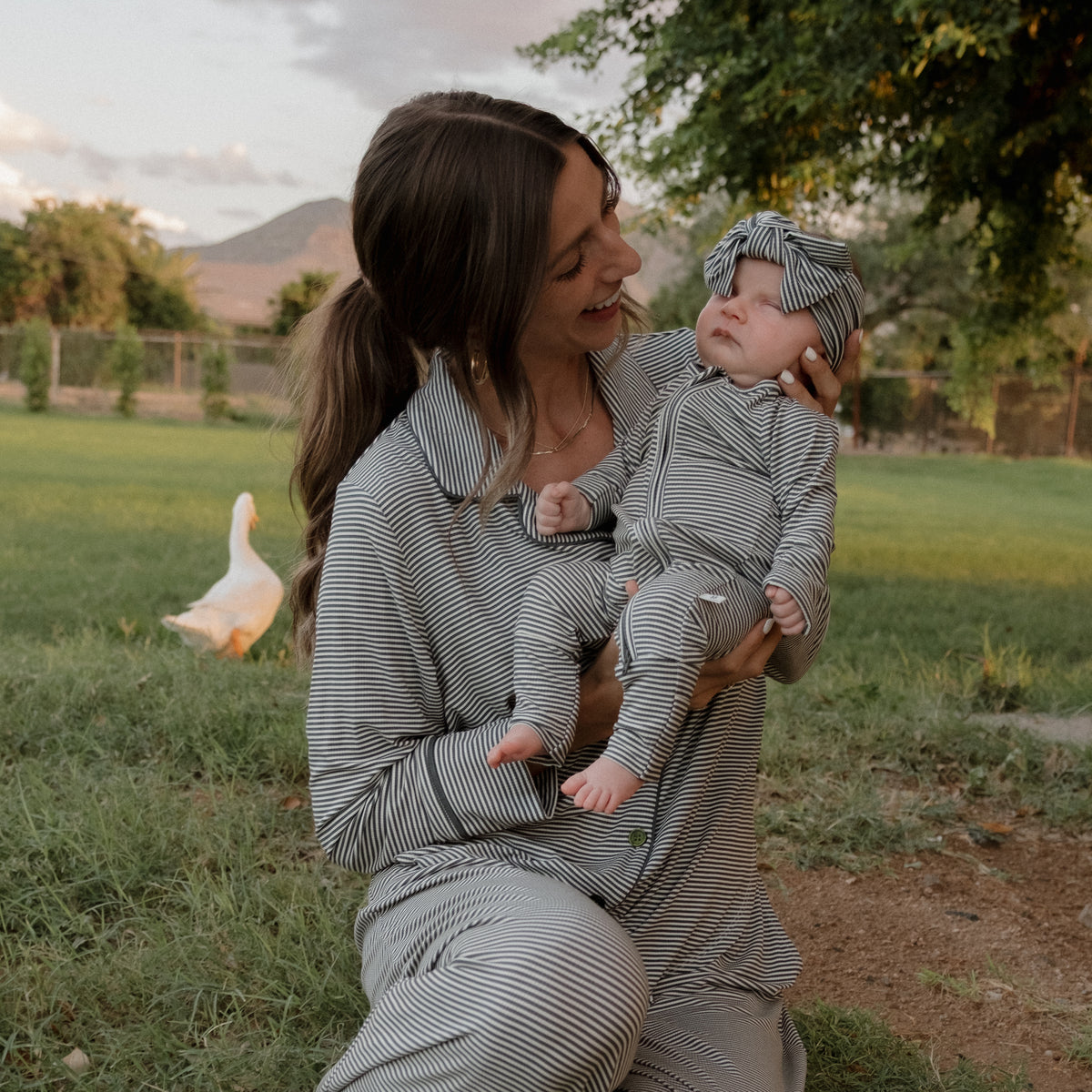 A woman wearing forever french baby’s Forest Stripe Ribbed Bamboo Zip Pajamas sits on the grass, holding a baby dressed in matching bamboo zip pajamas and a coordinating headband. In the background, a white duck wanders near trees and mountains. Both look happy and relaxed outdoors, enjoying the hypoallergenic comfort and softness of their forever french baby bamboo clothing.