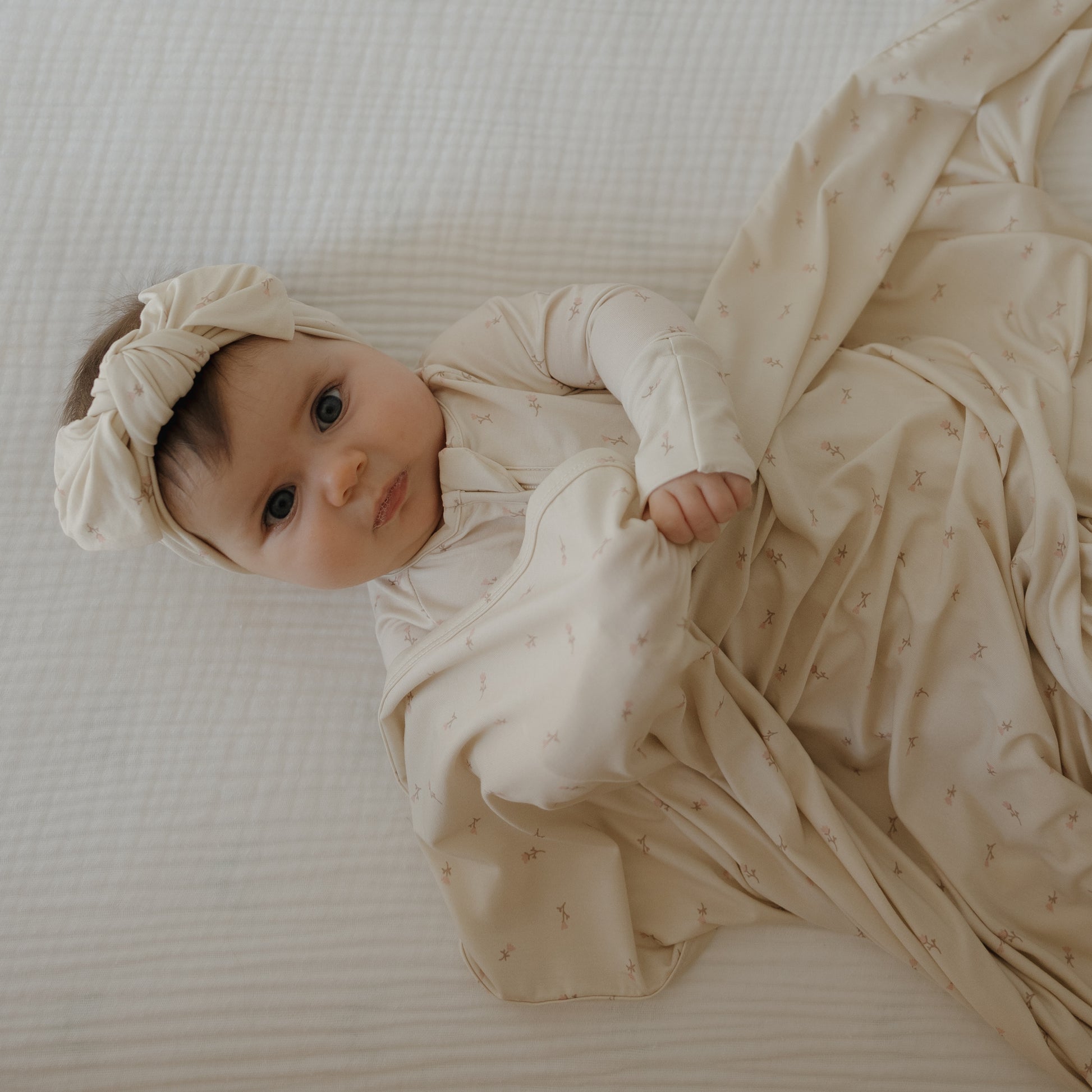A baby with blue eyes lies on a white bed, wearing forever french baby's Bamboo Zip Pajamas | Lucy, wrapped in a cream blanket and matching headband, looking up at the camera.