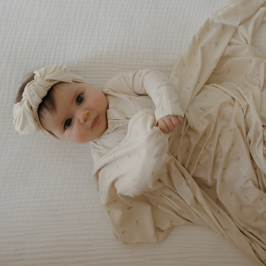A baby with blue eyes lies on a white bed, wearing forever french baby's Bamboo Zip Pajamas | Lucy, wrapped in a cream blanket and matching headband, looking up at the camera.