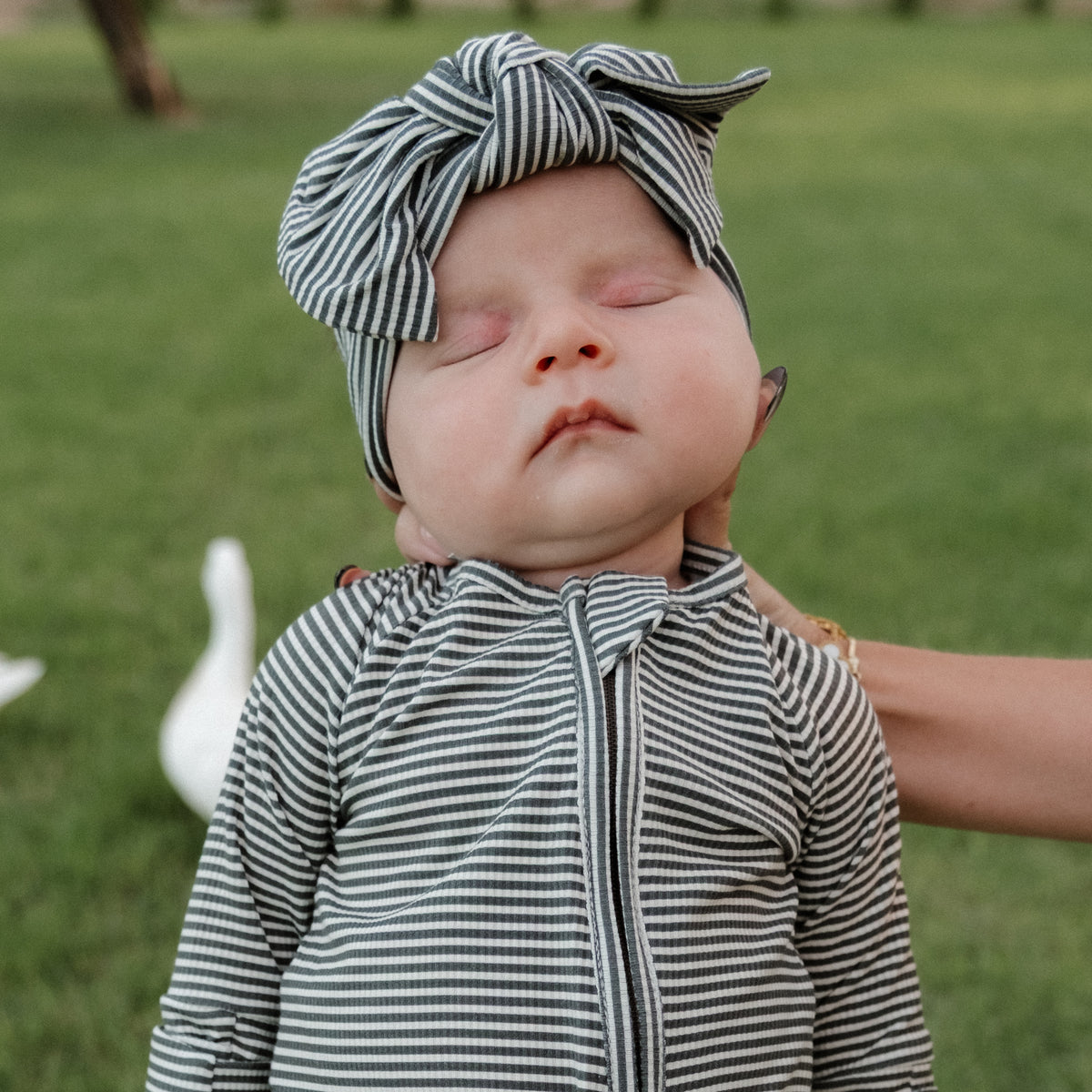 A baby dressed in forever french baby Bamboo Zip Pajamas | Forest Stripe Ribbed with a matching headband is held upright outdoors on green grass, eyes closed and looking peaceful. A white bird can be seen in the background.