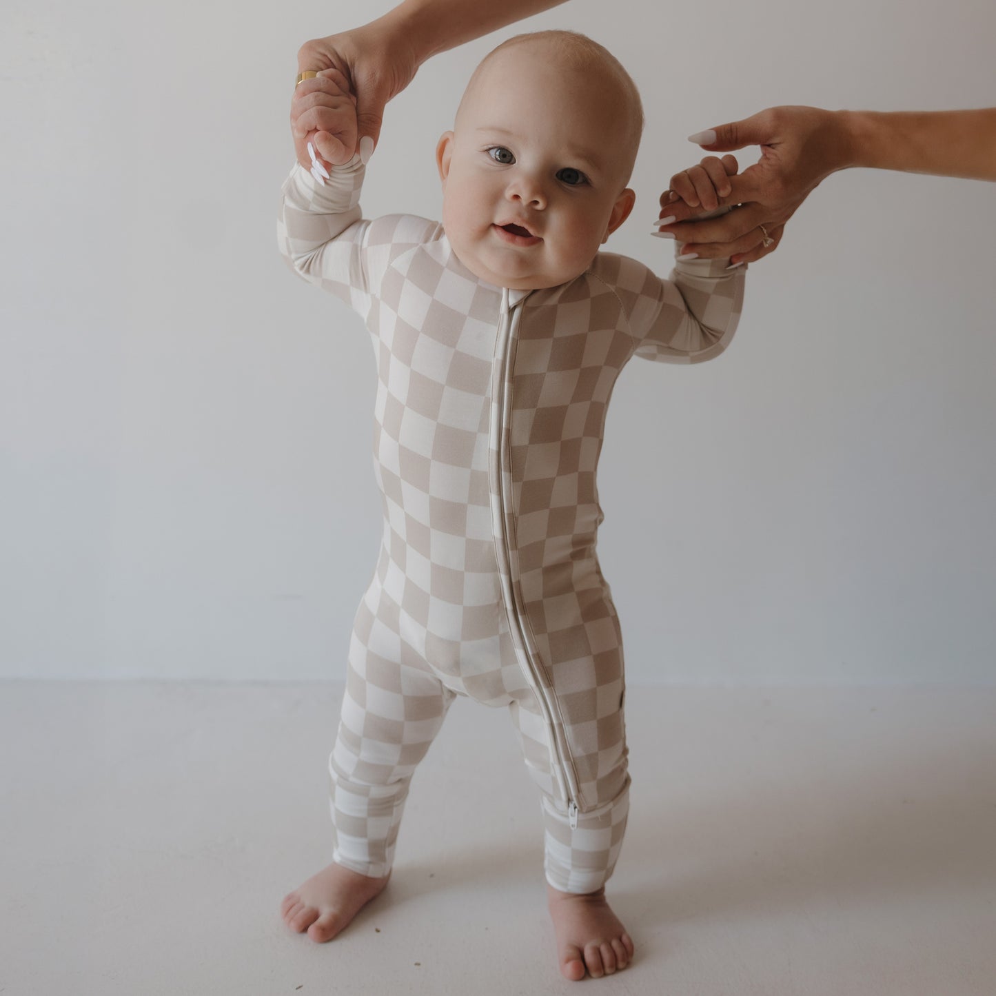 A baby in Forever French Baby Bamboo Zip Pajamas | Checkerboard stands supported by an adult’s hands, highlighting these hypoallergenic bamboo pajamas against a plain light background.