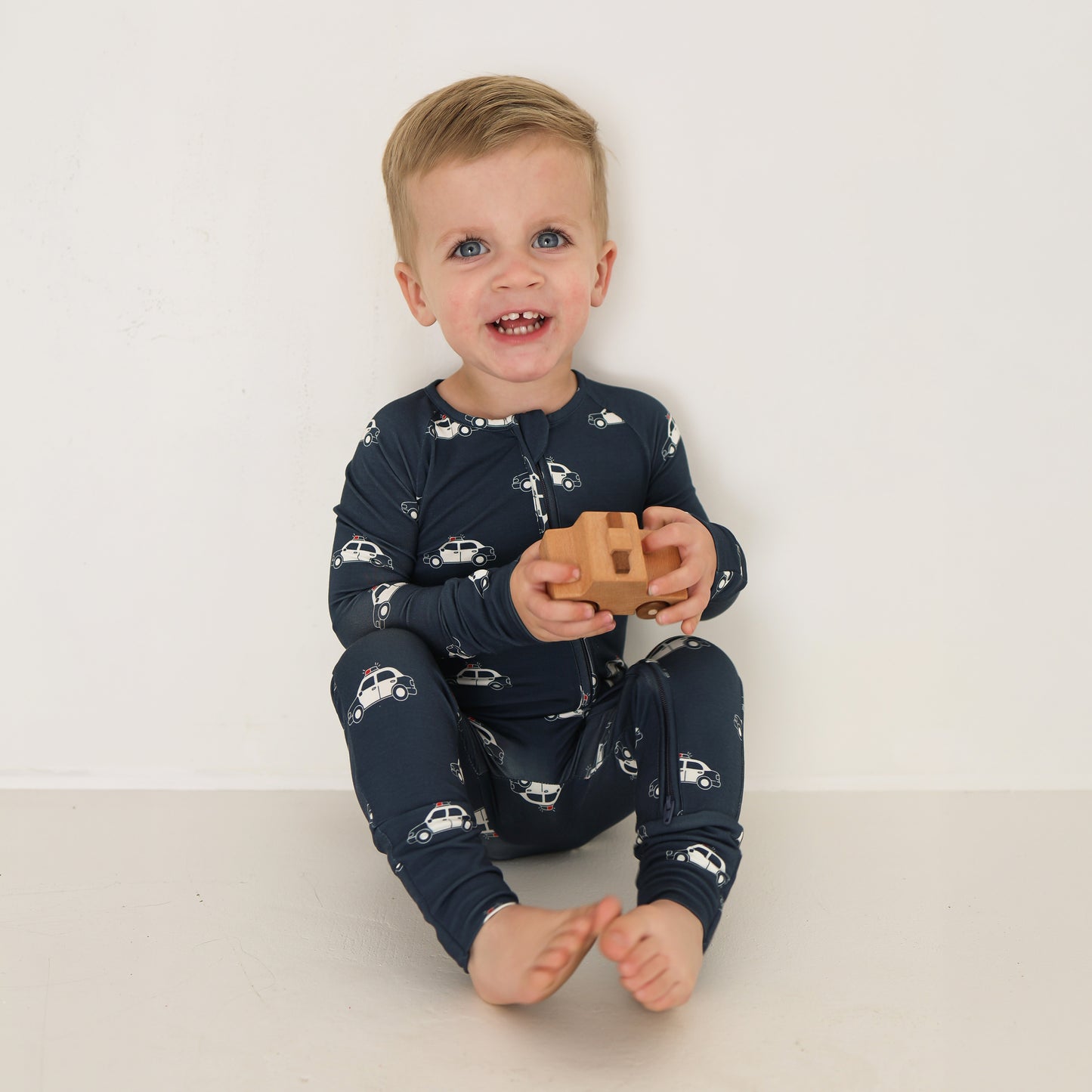 A blond toddler, smiling and holding a wooden toy car, sits against a white wall wearing forever french baby Bamboo Zip Pajamas | Police Cruiser.