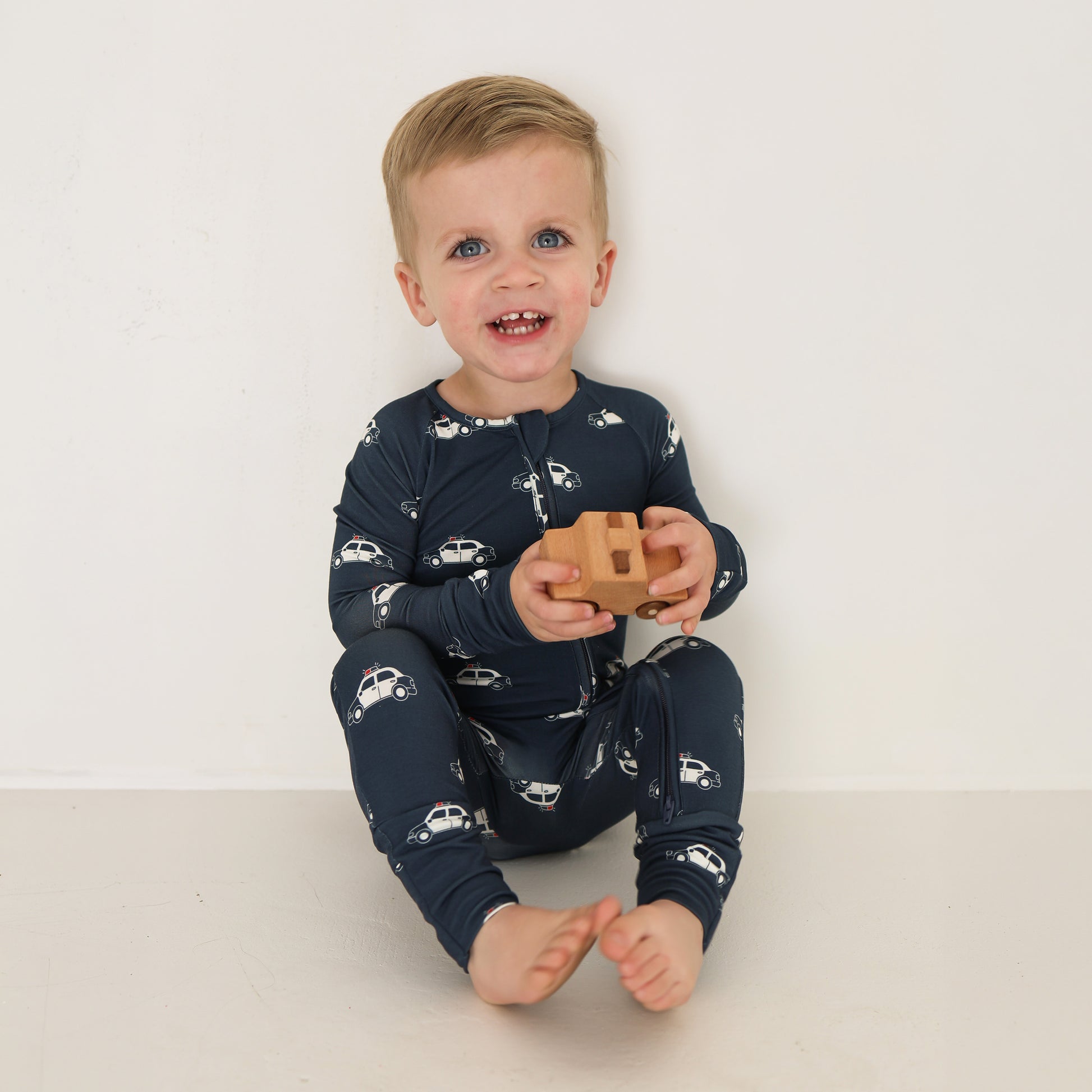 A blond toddler, smiling and holding a wooden toy car, sits against a white wall wearing forever french baby Bamboo Zip Pajamas | Police Cruiser.