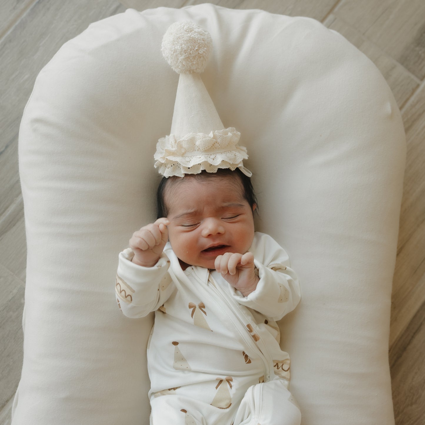 A newborn baby wearing the forever french baby Bamboo Zip Pajamas | Birthday Girl and a small party hat lies on a soft white cushion with eyes closed and fists raised, set against a wooden floor background.