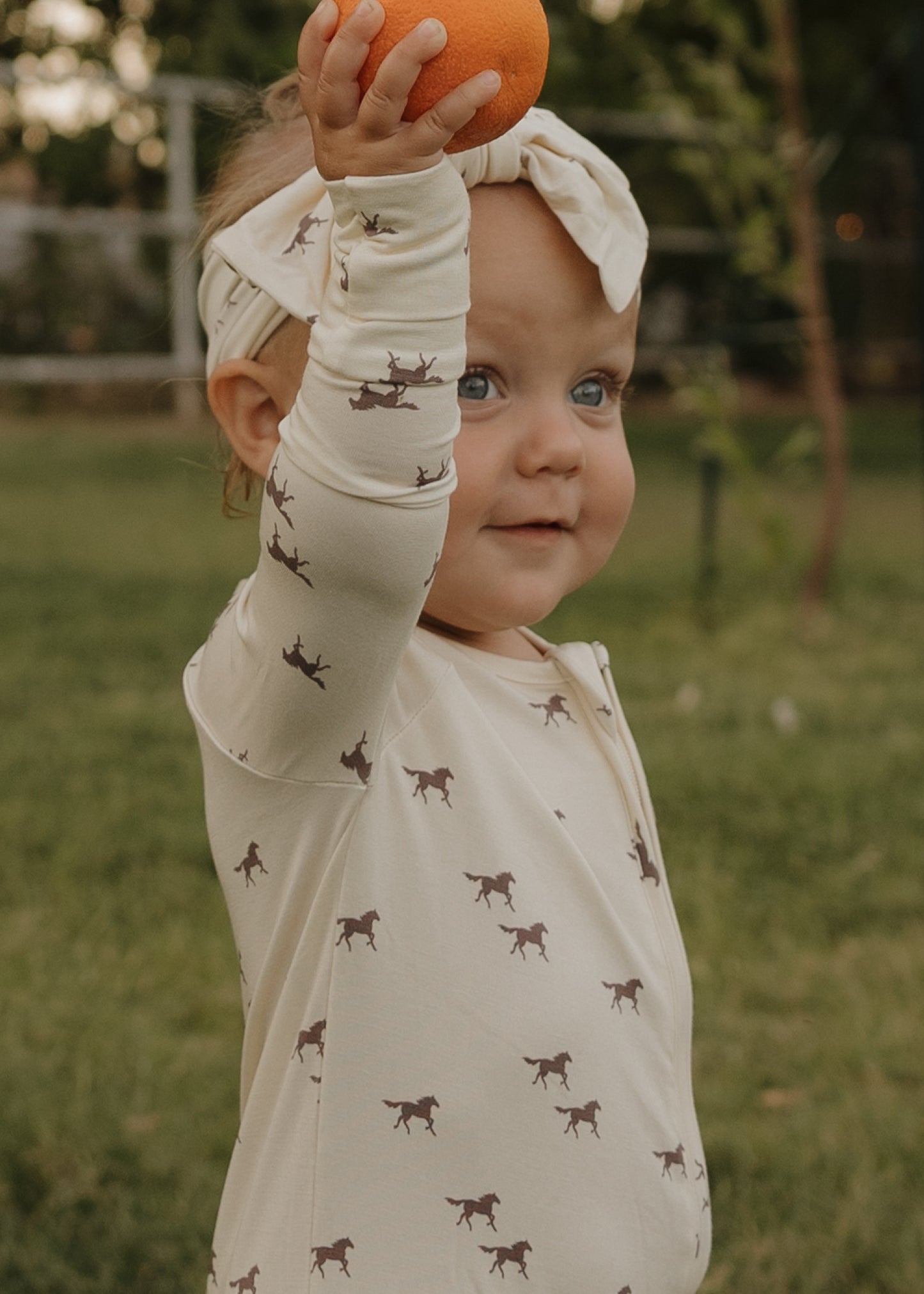 A smiling toddler wearing forever french baby Bamboo Zip Pajamas | Wild Horses—cream pajamas with brown horse prints and a matching headband—holds up an orange while standing on grass.
