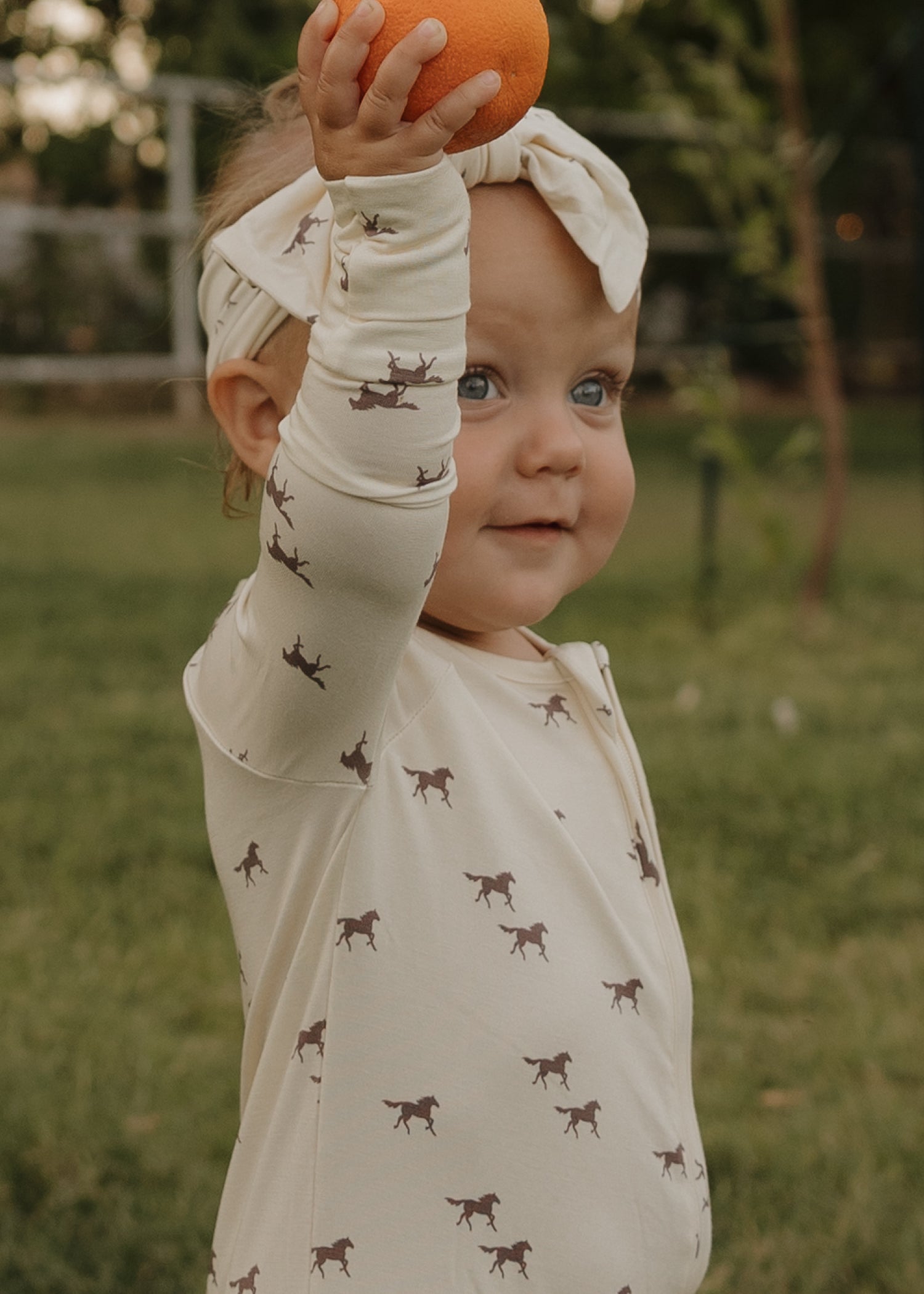 A smiling toddler wearing forever french baby Bamboo Zip Pajamas | Wild Horses—cream pajamas with brown horse prints and a matching headband—holds up an orange while standing on grass.