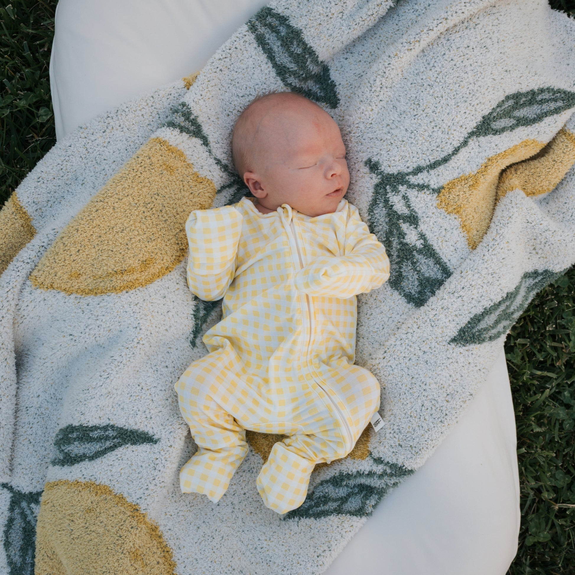 A newborn in forever french baby Bamboo Zip Pajamas | Citron, featuring a yellow and white check pattern, sleeps on a lemon and green leaf blanket atop a white outdoor cushion.