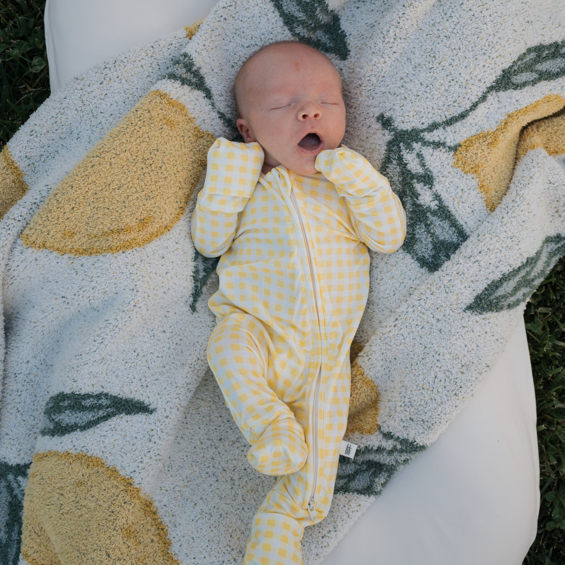 A baby in forever french baby’s Bamboo Zip Pajamas | Citron yawns, eyes closed and hands near their face, lying on a white and yellow lemon-patterned blanket.