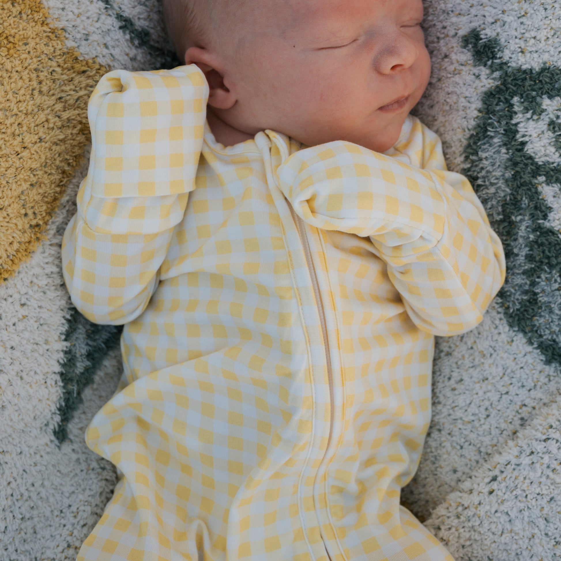A newborn sleeps peacefully in forever french baby's Bamboo Zip Pajamas | Citron—a yellow and white checkered onesie with built-in mittens—resting on a soft, textured blanket.