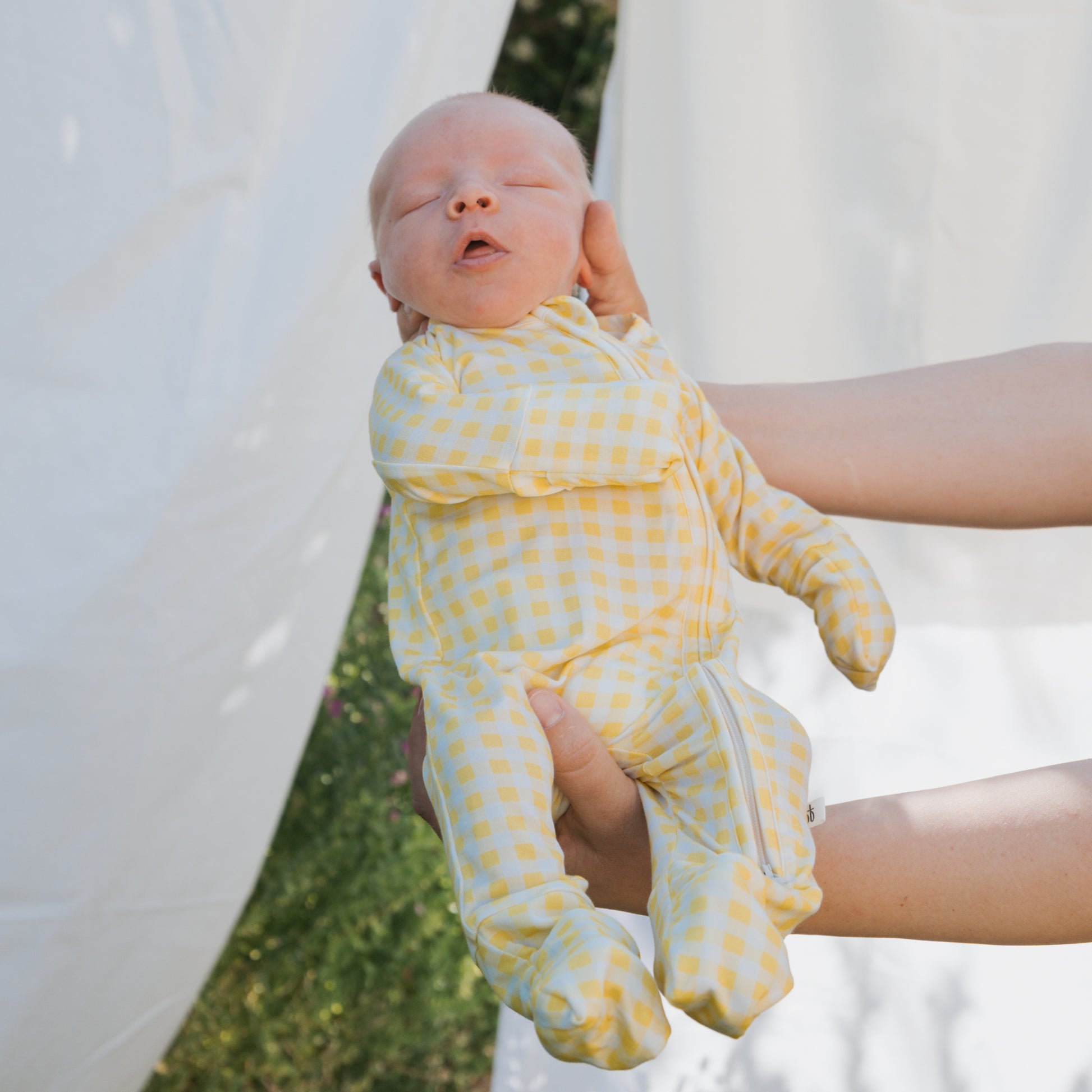 A newborn in forever french baby's hypoallergenic Bamboo Zip Pajamas in the Citron print is gently held outdoors by two adult hands, with white fabric and greenery in the background.