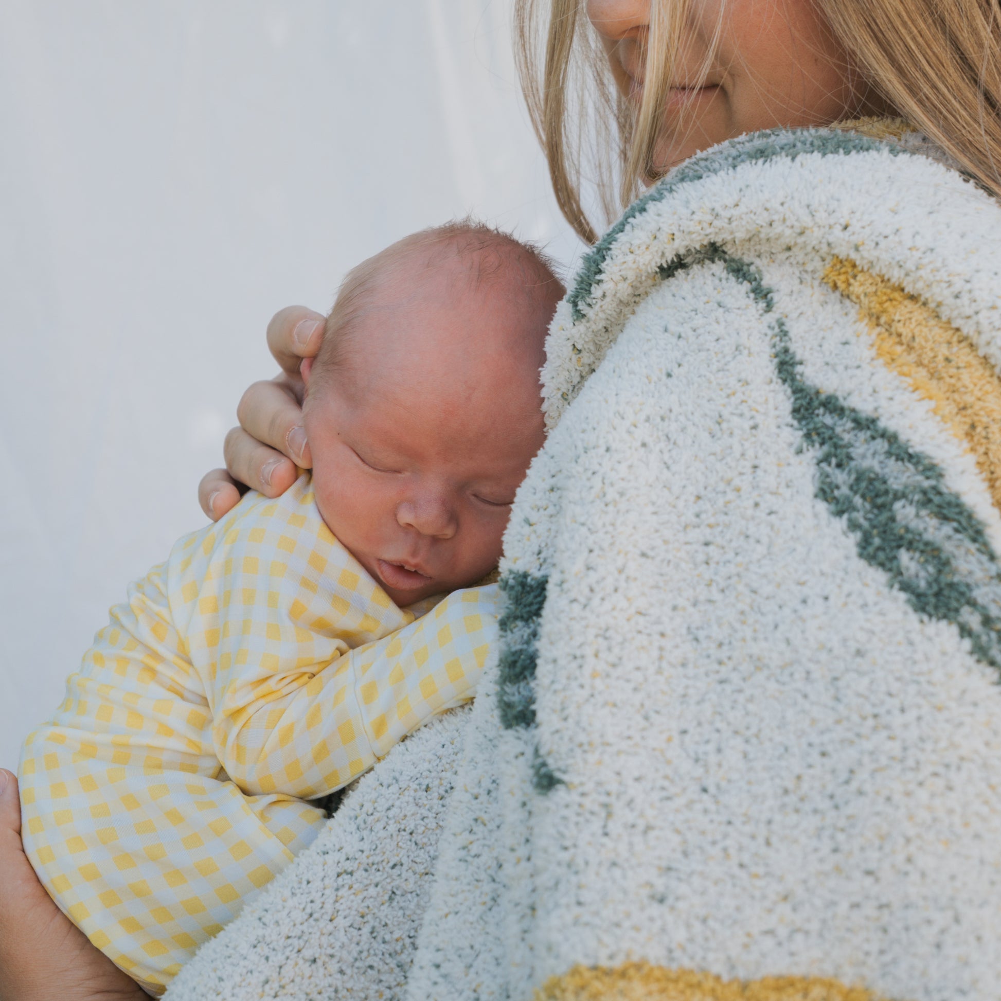 A woman gently holds a sleeping newborn dressed in forever french baby’s Bamboo Zip Pajamas | Citron, wrapped in a cozy blanket. The baby's head rests on her shoulder, highlighting the comfort of these hypoallergenic pajamas.