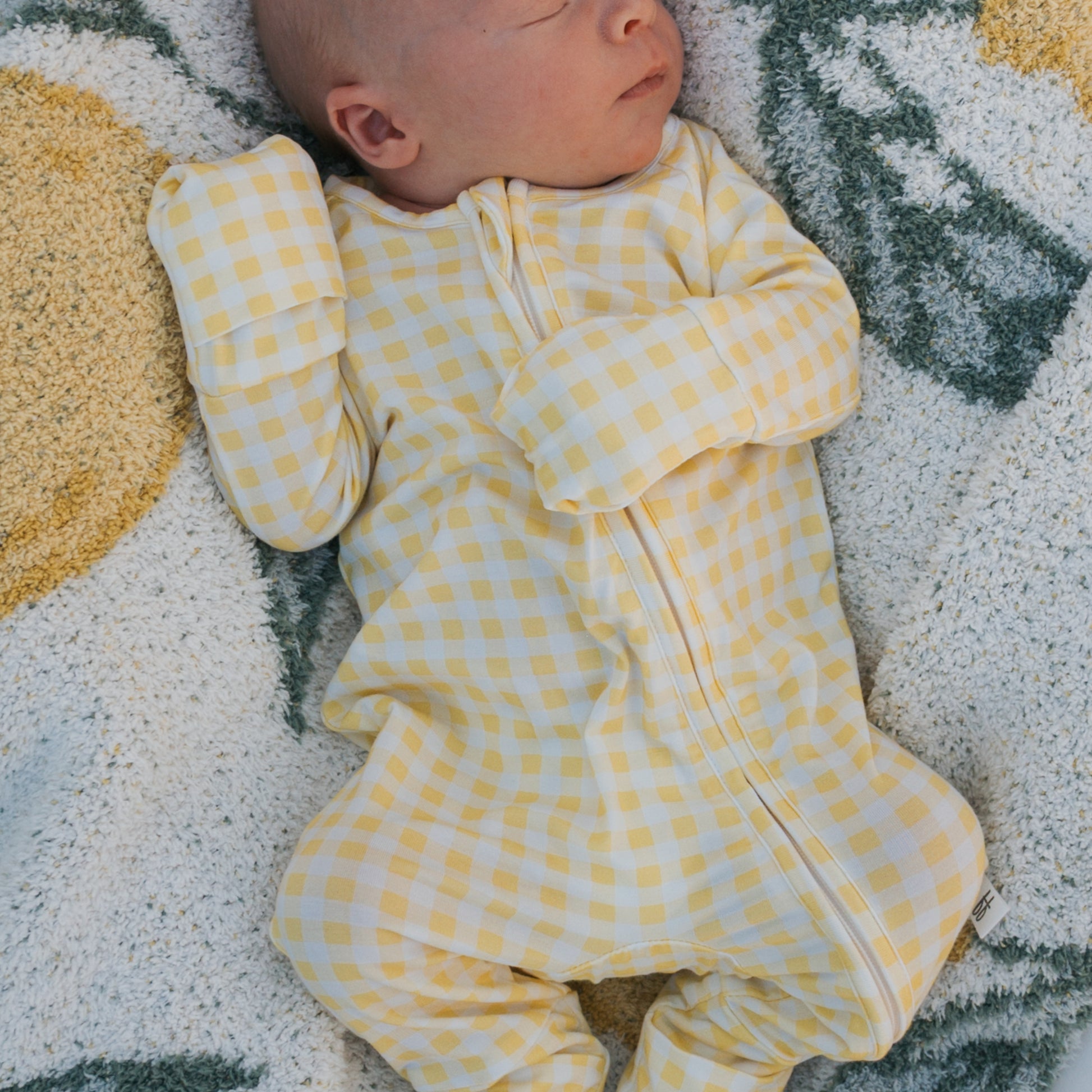 A newborn rests peacefully with eyes closed, wearing forever french baby's Bamboo Zip Pajamas in Citron—a yellow and white checkered, breathable onesie with covered hands—lying on a soft, patterned blanket.