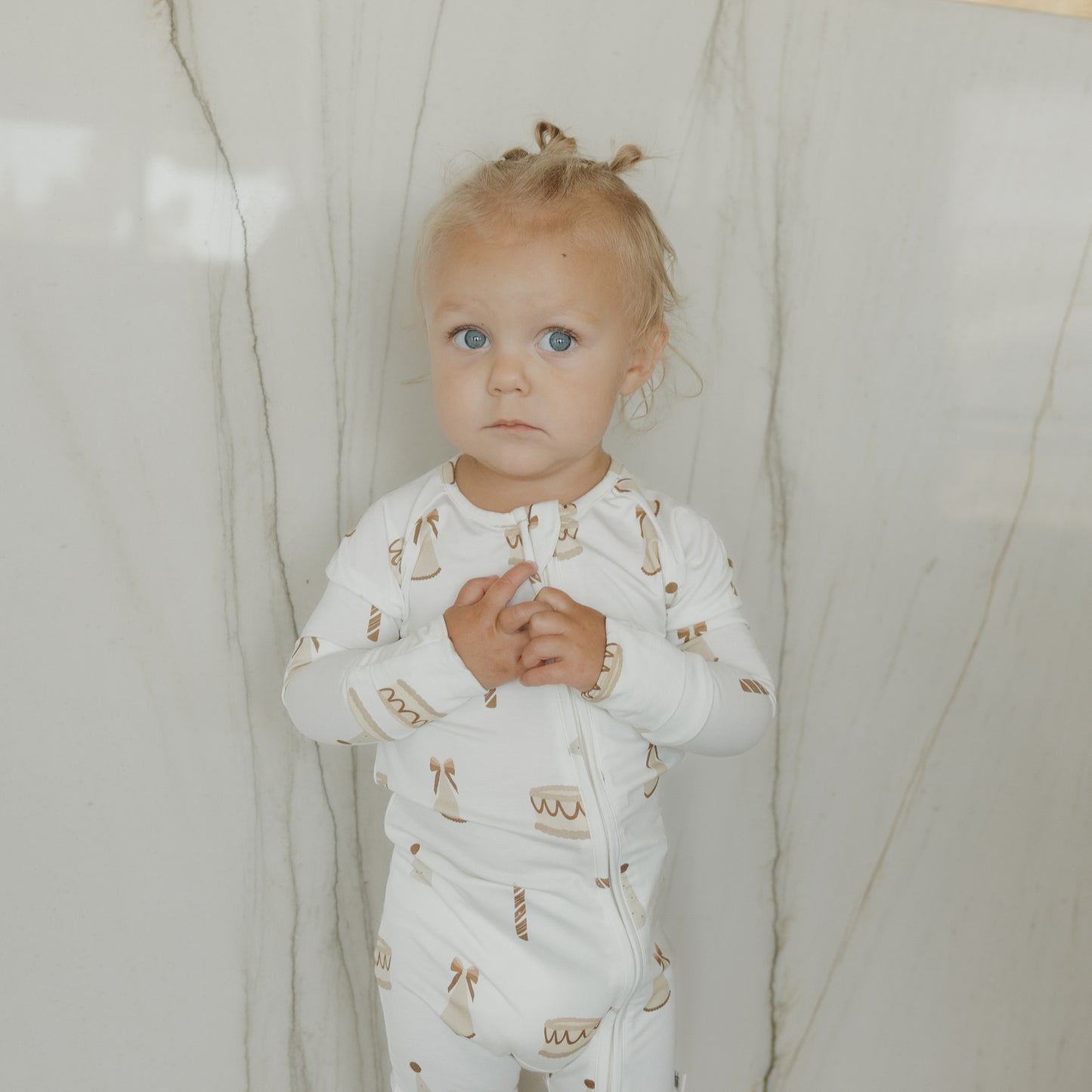 A toddler with blonde hair in small buns, wearing forever french baby Bamboo Zip Pajamas | Birthday Girl—white hypoallergenic baby clothing adorned with brown cake and candle designs—stands against a light-colored marble wall, looking at the camera.