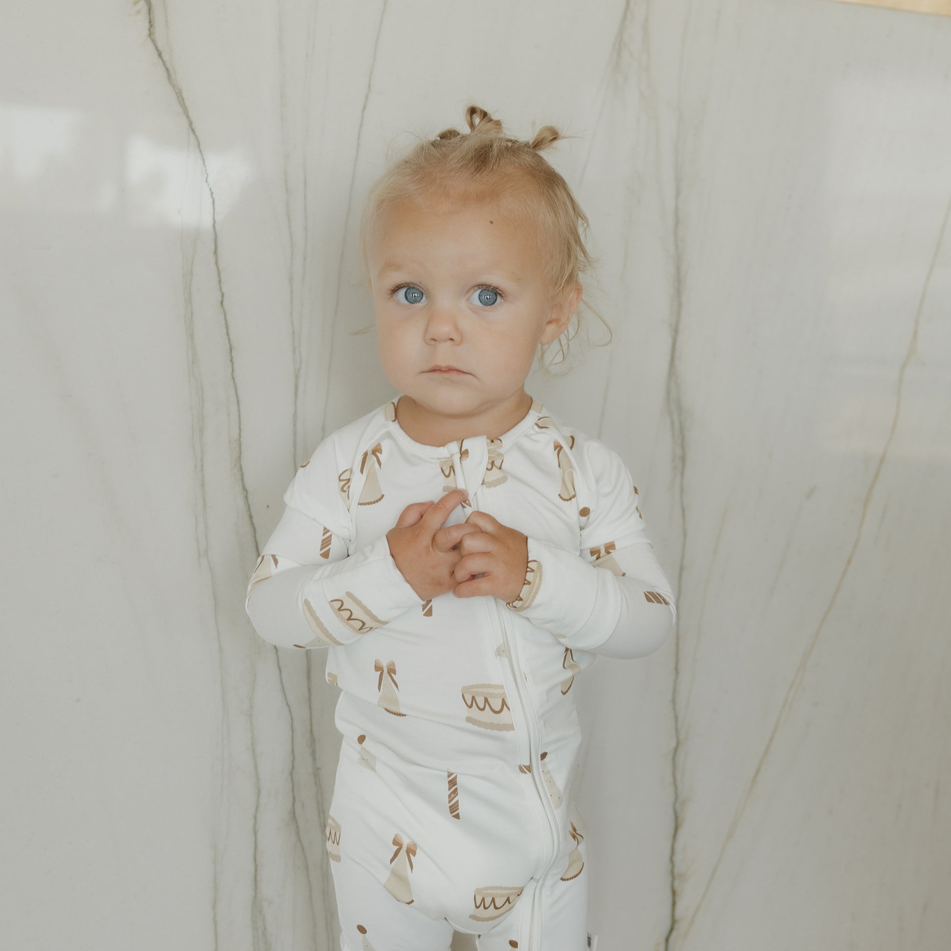 A toddler with blonde hair in small buns, wearing forever french baby Bamboo Zip Pajamas | Birthday Girl—white hypoallergenic baby clothing adorned with brown cake and candle designs—stands against a light-colored marble wall, looking at the camera.