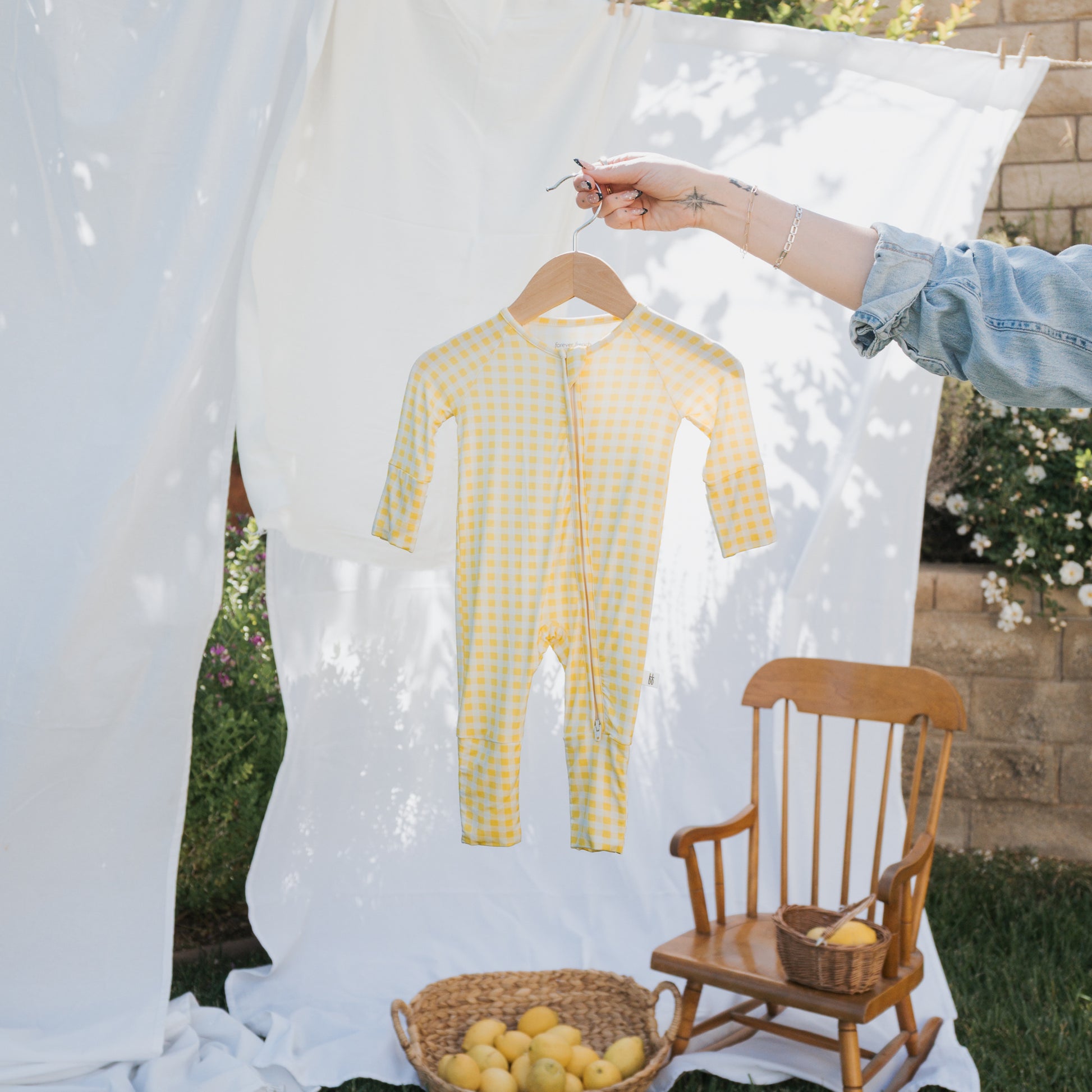 A person holds up the forever french baby Bamboo Zip Pajamas | Citron, a yellow and white checkered, breathable baby onesie, on a hanger outdoors. In the background are white sheets, a wooden rocking chair, and a basket with yellow apples.