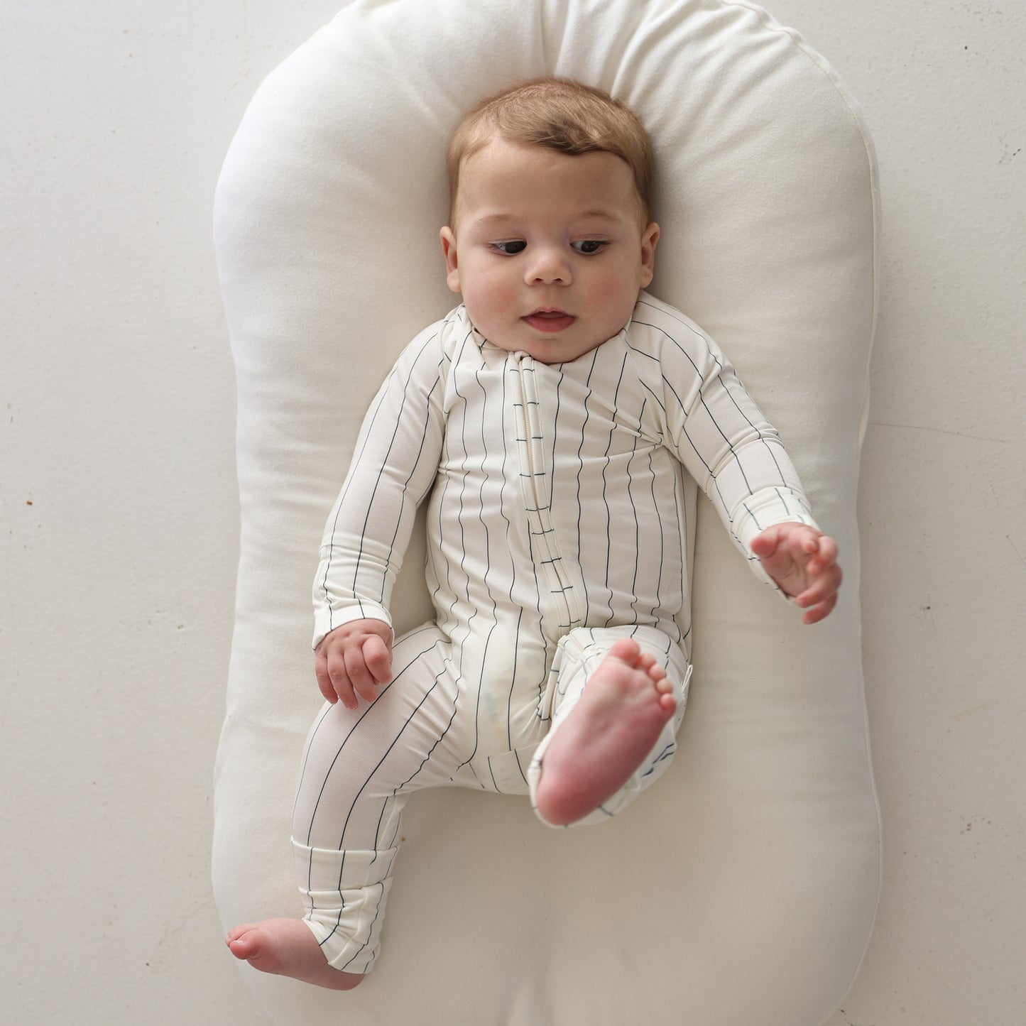 A baby in forever french baby's Bamboo Zip Pajamas | Jack—a white, long-sleeve bodysuit with black stripes—lies on a soft white cushion, looking to the side with one leg raised and one hand extended.