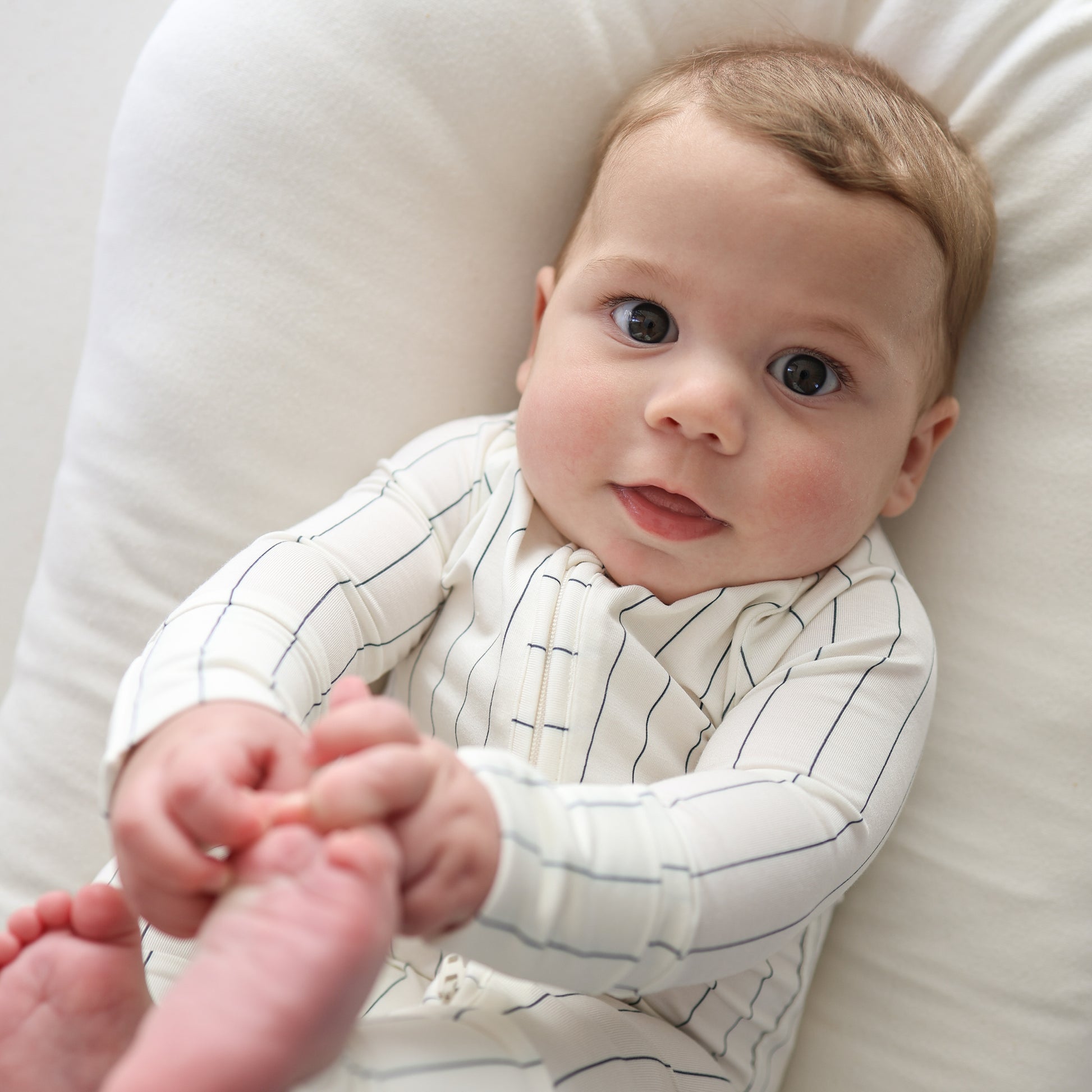 A baby in forever french baby Bamboo Zip Pajamas | Jack relaxes on a soft cushion, gazing upward with wide eyes and a gentle smile while grabbing their feet with both hands.