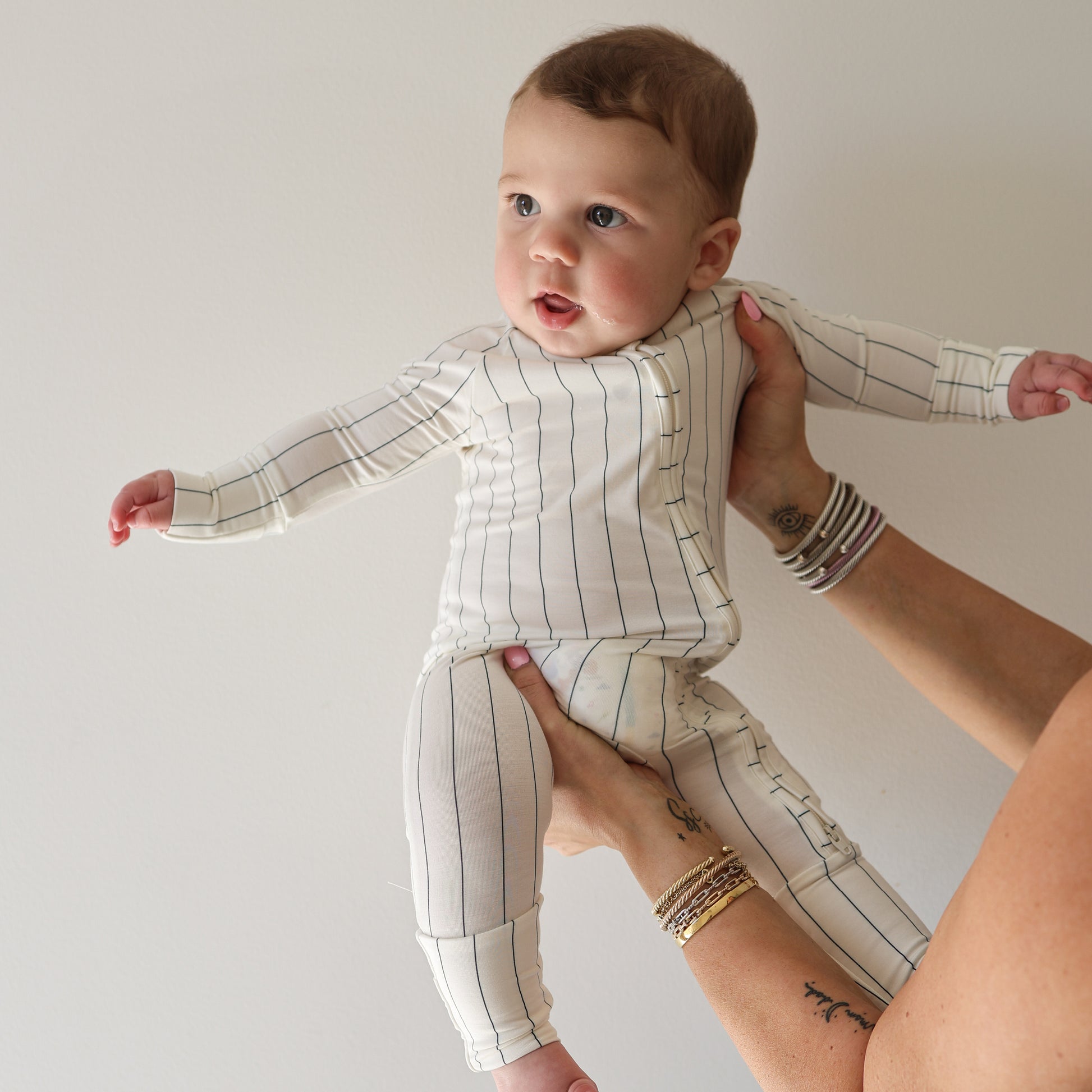 A baby in forever french baby's Bamboo Zip Pajamas | Jack—a white, long-sleeve outfit with thin black stripes—is lifted by an adult wearing bracelets, set against a plain light background.