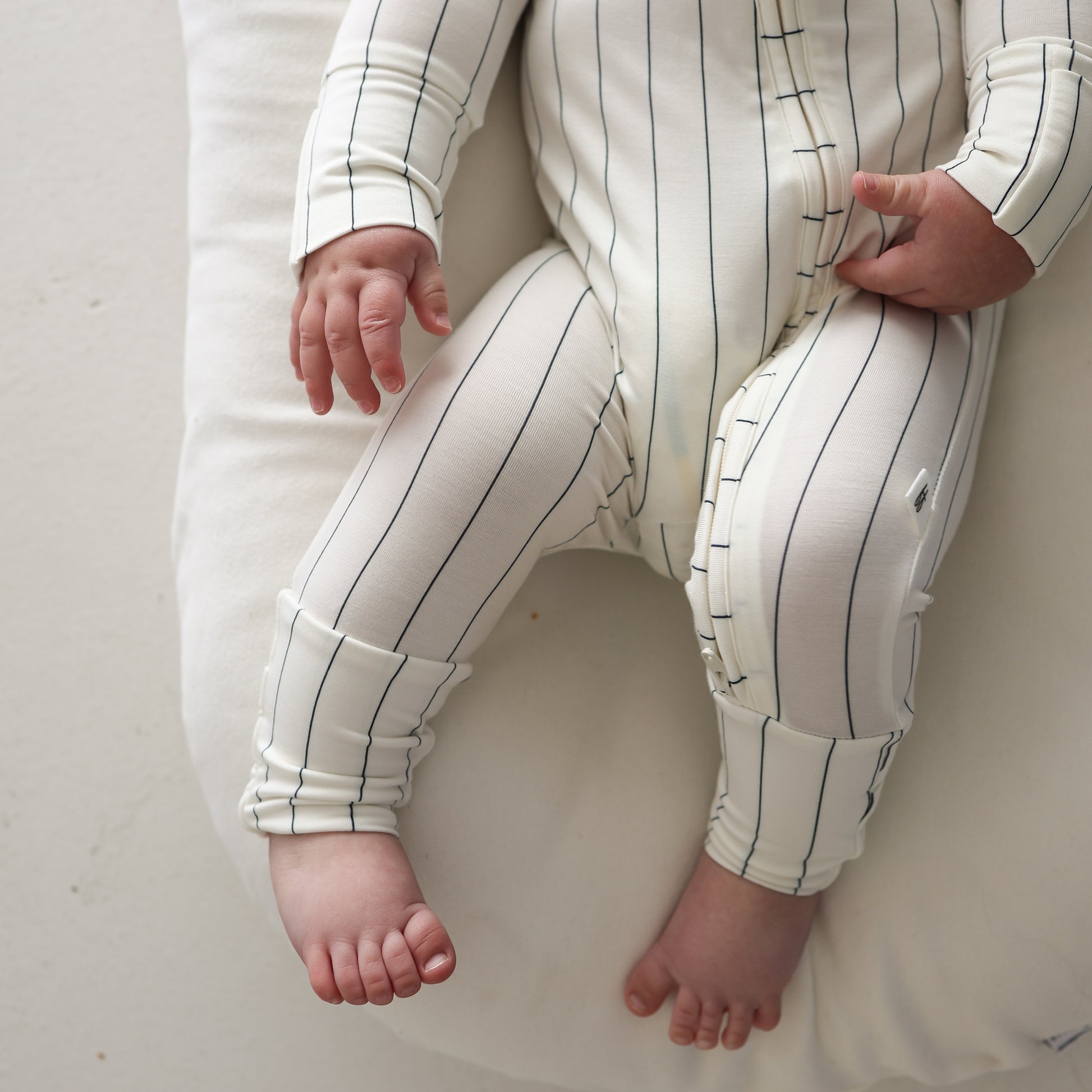 A baby sits on a soft white cushion, wearing forever french baby Bamboo Zip Pajamas | Jack—a breathable, hypoallergenic white onesie with thin black stripes. Only the baby's torso, arms, and legs are visible.