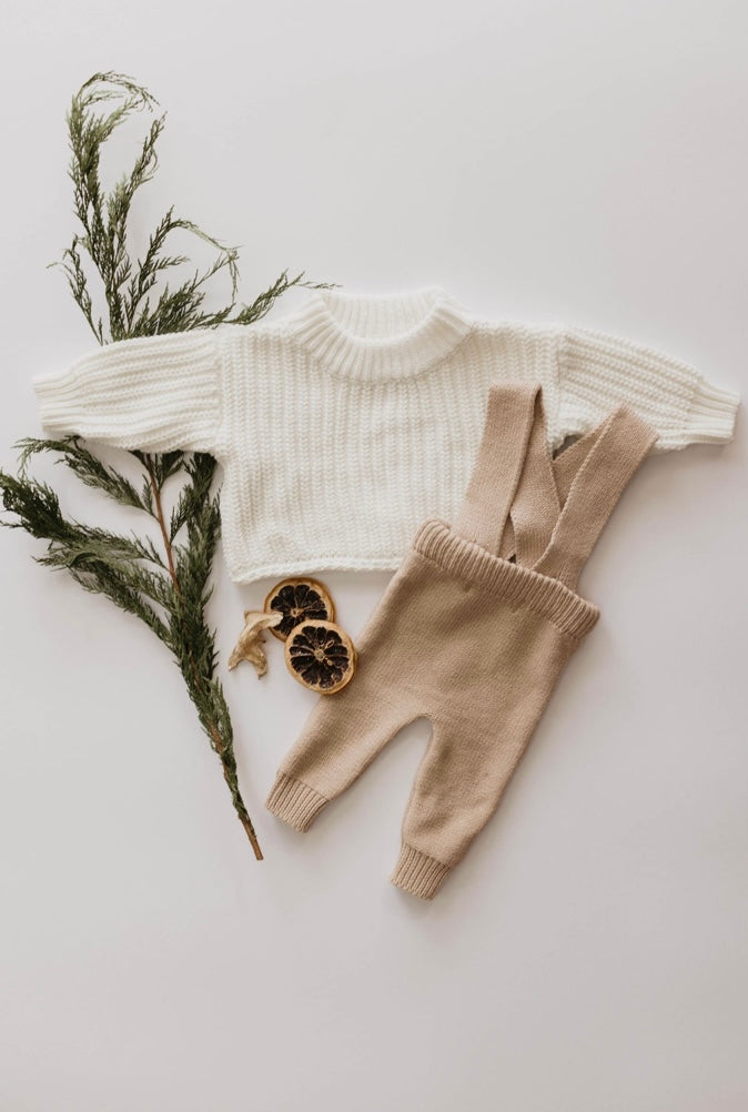 A flat lay of Forever French's Knit Suspenders in a cream-colored sweater and beige pants, accompanied by green pine branches and dried orange slices, evokes the cozy charm of cooler weather. Arranged on a light background, this scene captures the perfect seasonal vibe.