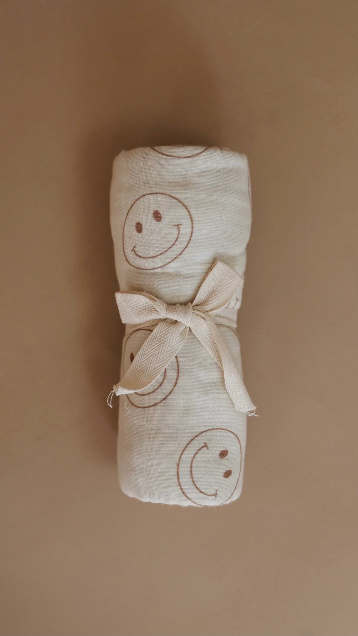 The Just Smile Ivory muslin swaddle from forever french baby is neatly rolled up and secured with a cream-colored ribbon. The blanket features a pattern of smiling faces printed in brown, all set against a simple beige background.